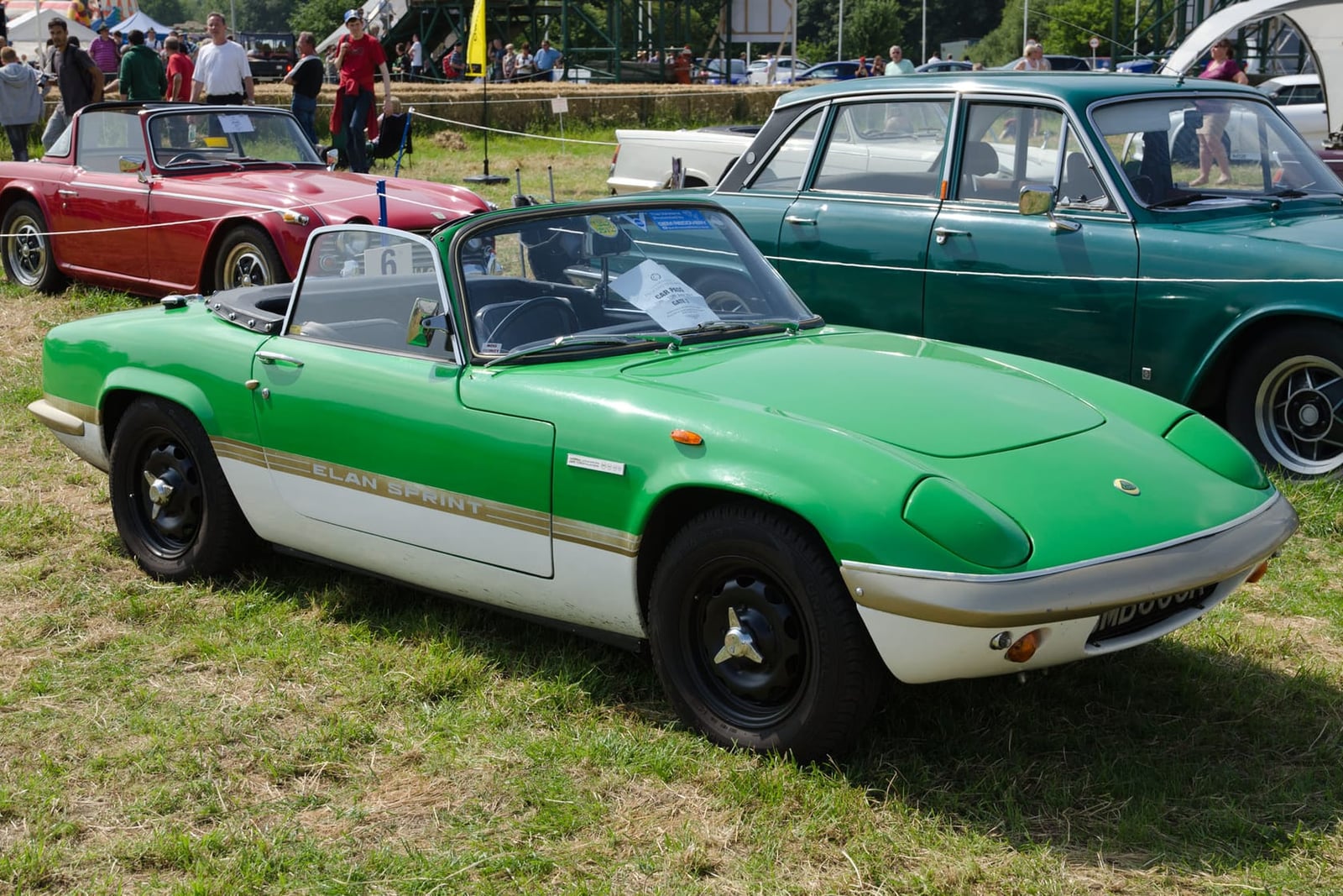A green and white 1972 Lotus Elan Sprint with a UK registration on a show field, a saloon behind it