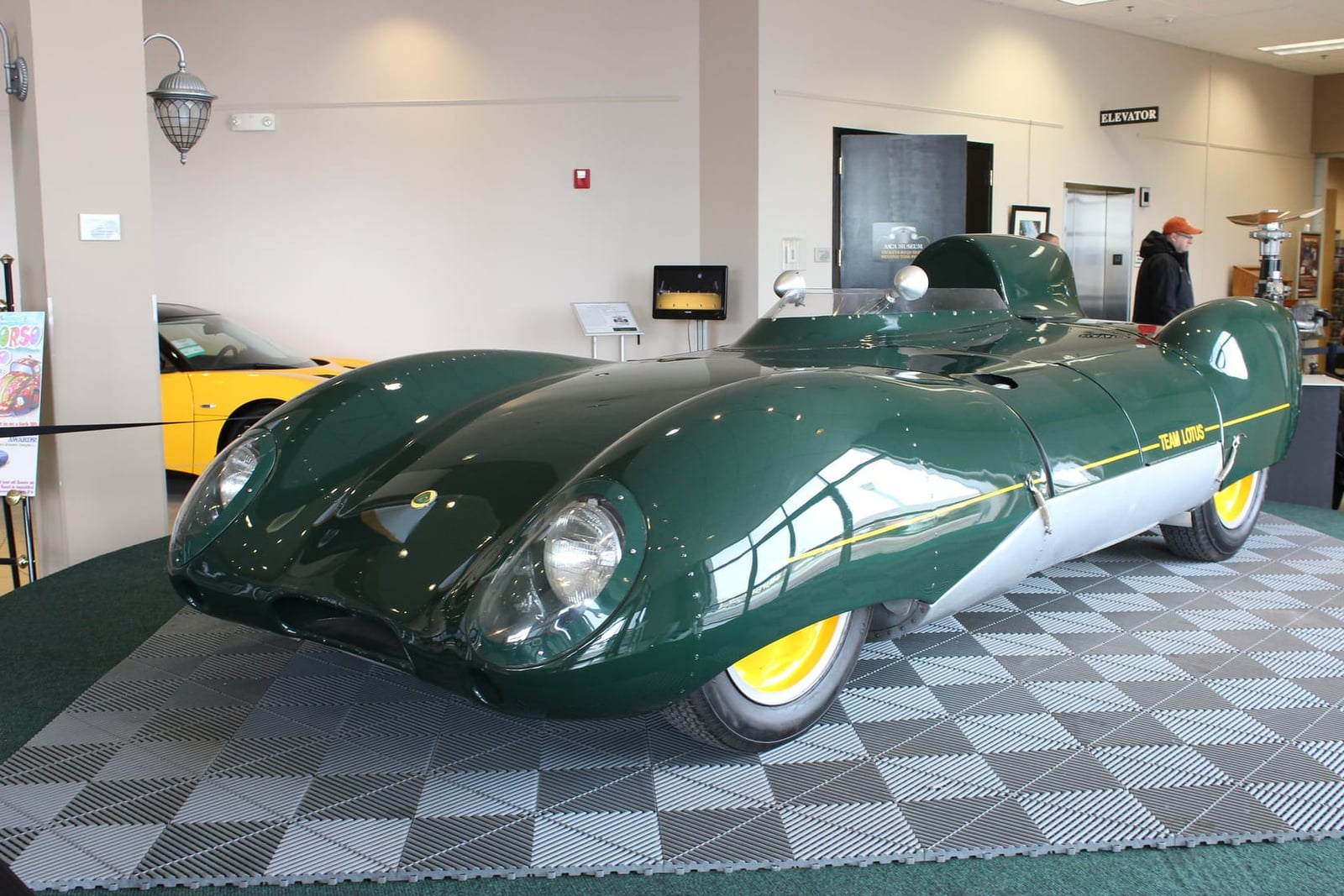 A green Lotus Eleven Series 2 Le Mans displayed indoors on a checkered floor, front three-quarter view