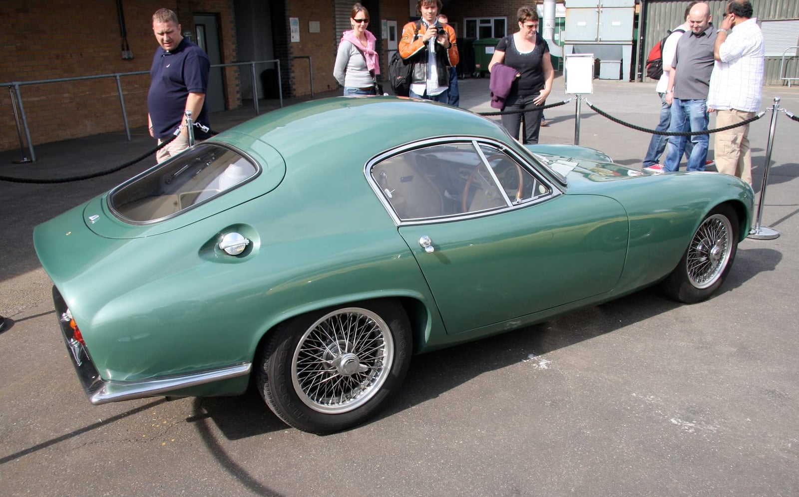 A green Lotus Elite parked on tarmac at a classic car gathering, rear three-quarter view showing the fastback roofline