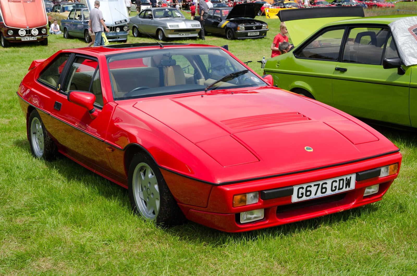 A red 1989 Lotus Excel SE parked on grass at a classic car show, front three-quarter view, UK registration G676 GDW