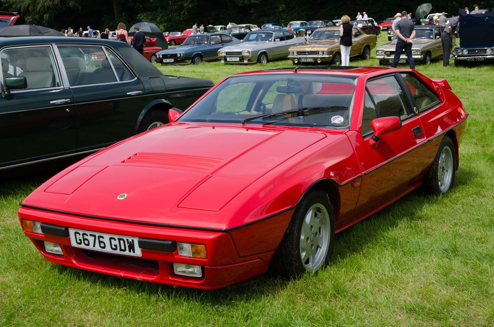 A red 1989 Lotus Excel parked on grass among other classic cars, front view
