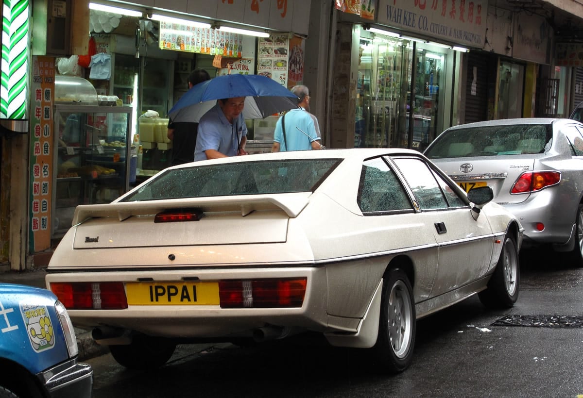 A white Lotus Excel parked on a busy shopping street in Hong Kong, rear three-quarter view