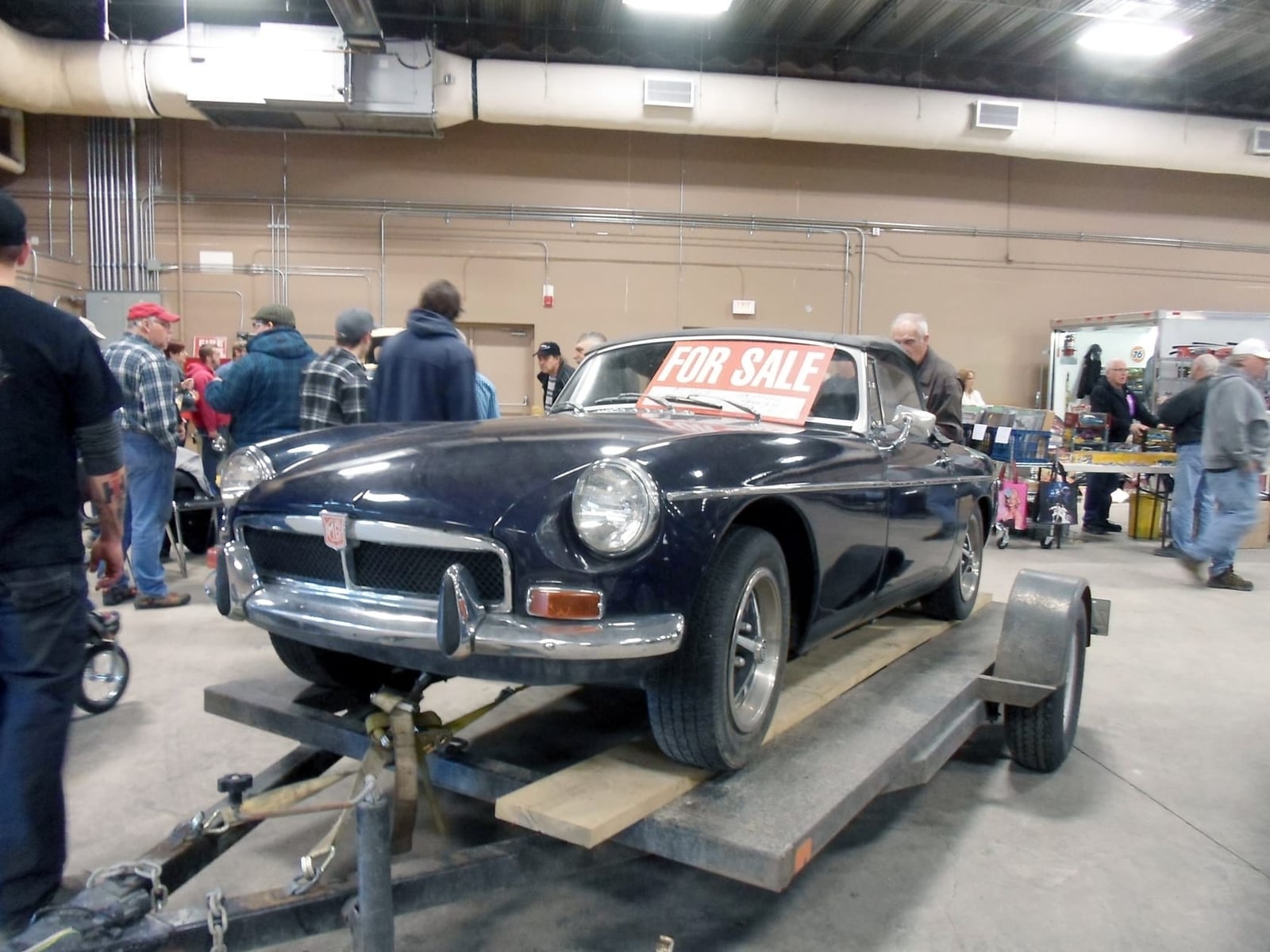 A dark blue chrome-bumper MG MGB roadster on a trailer with a For Sale sign in its windscreen at an indoor car show