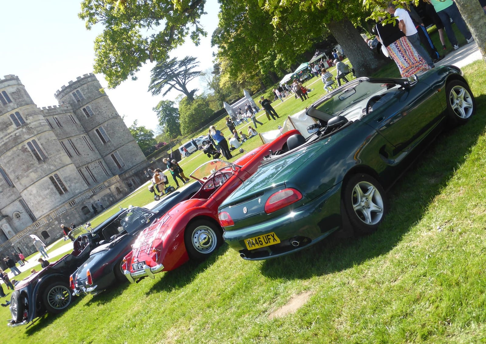 A dark green MGF parked with older MGs on the grass in front of Lulworth Castle