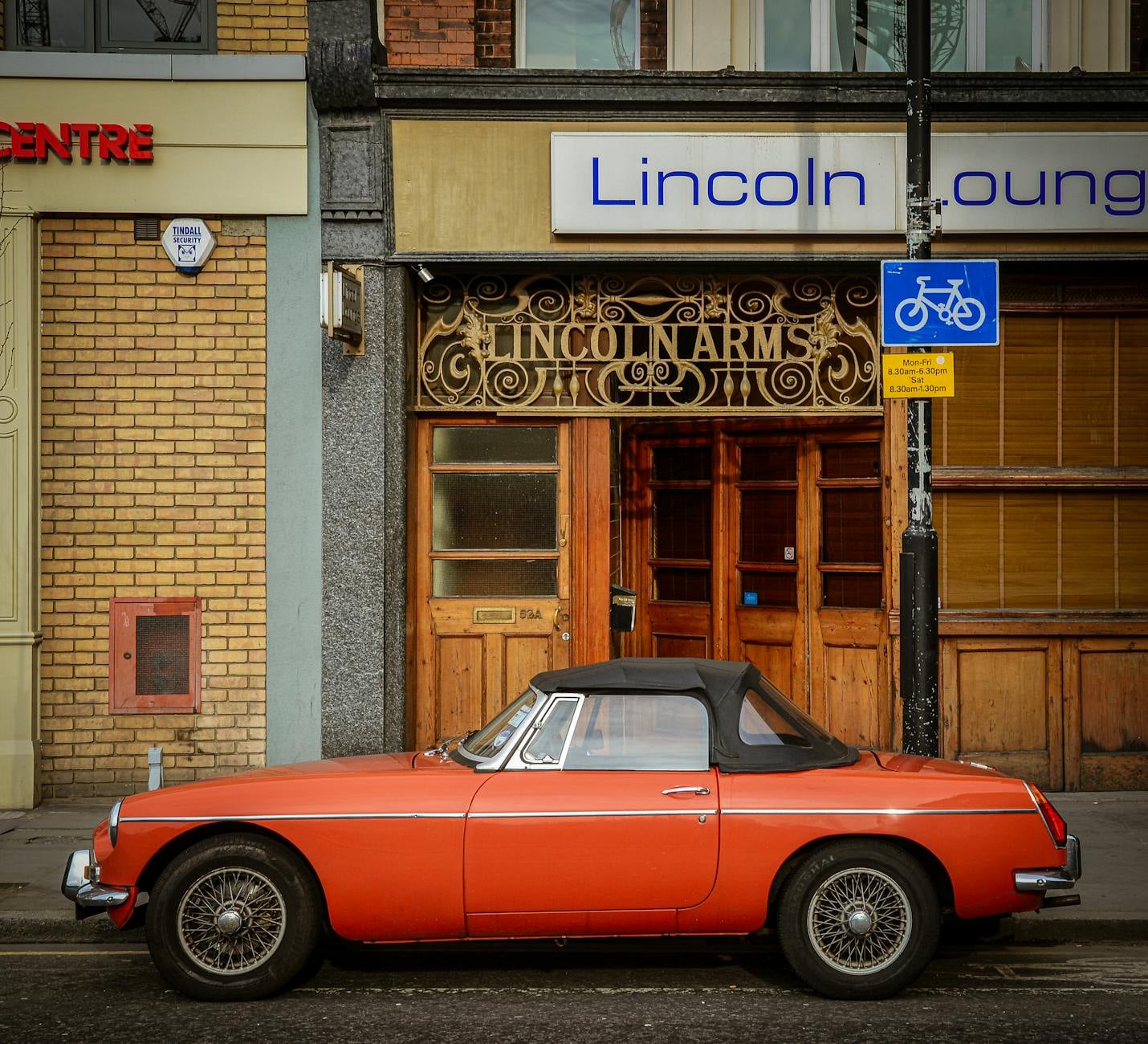 An orange MG MGB roadster with chrome wire wheels, side view parked outside a town pub