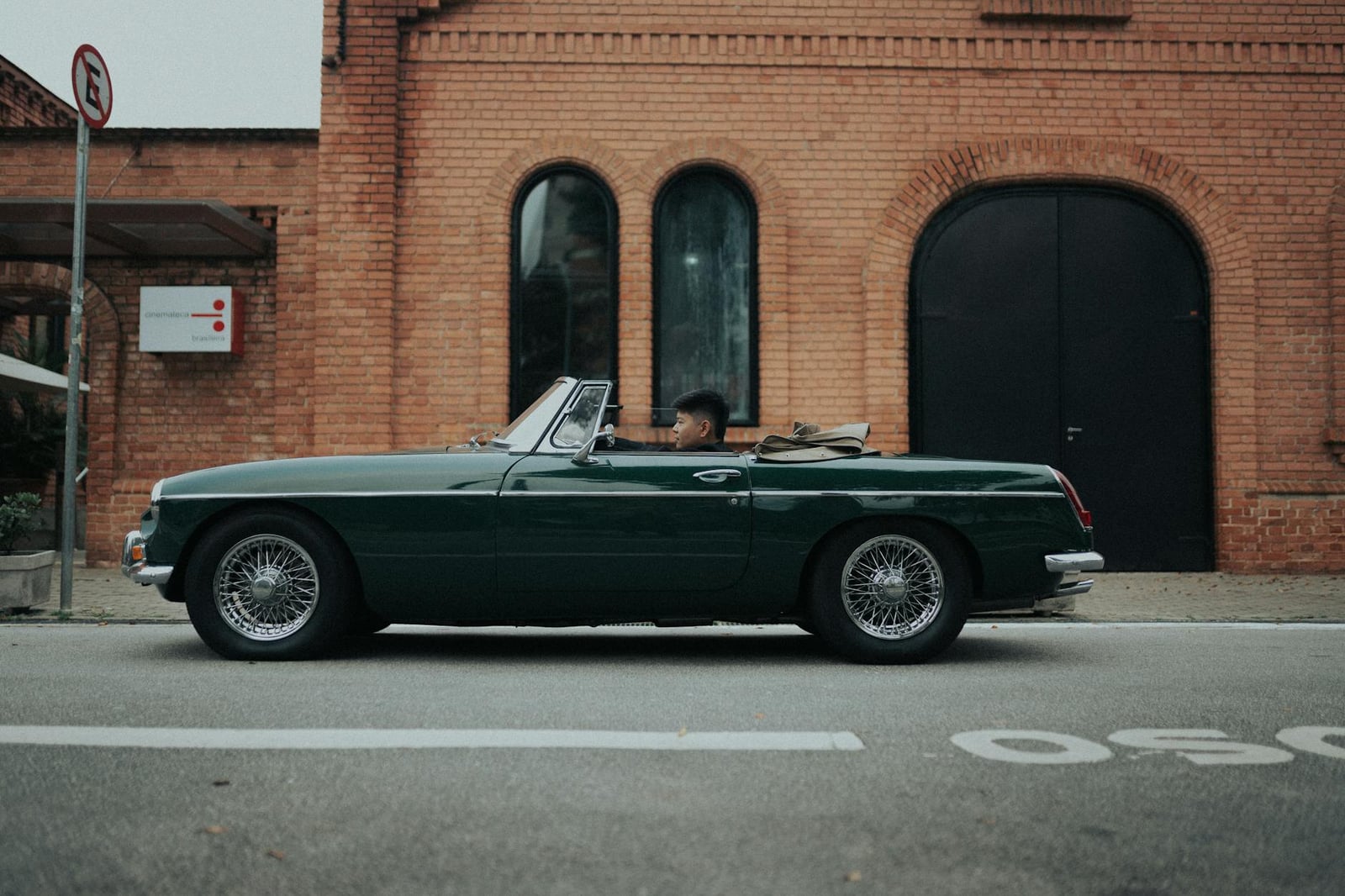 A British Racing Green MG MGB roadster with chrome wire wheels, side profile against a red-brick warehouse