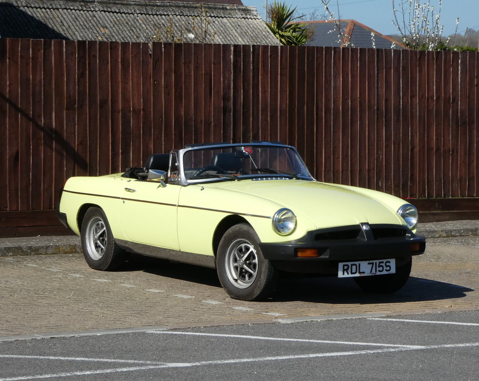A primrose yellow MG MGB roadster parked by a wooden fence, front three-quarter view