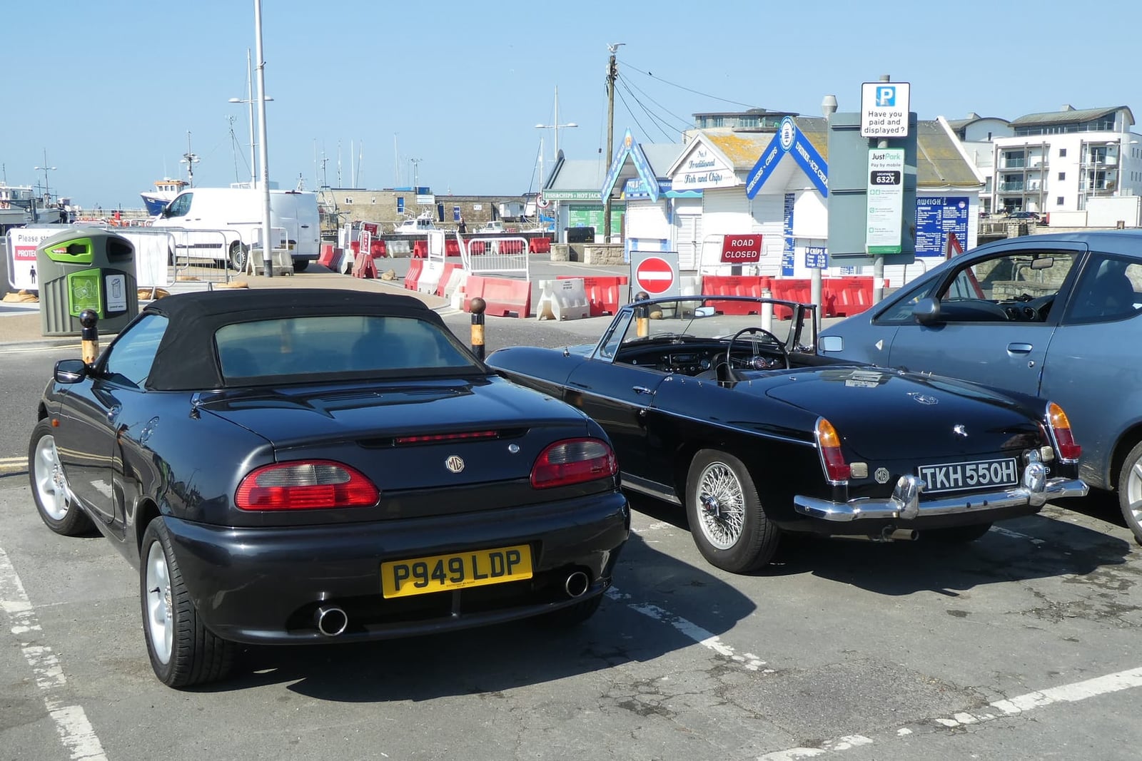 A black MGF beside a chrome-bumper MGB roadster at a harbour