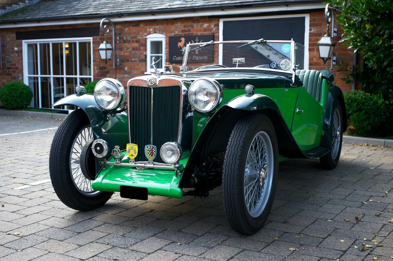A green MG TC, an upright pre-war-shaped sports car, parked by a brick house
