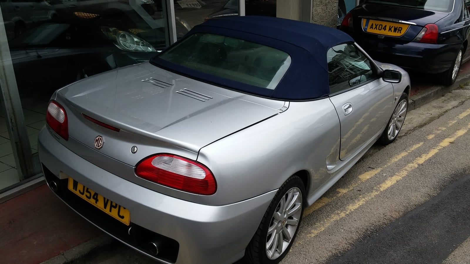 The rear of a silver 2004 MG TF 160 with its hood up and a UK registration, parked at a kerb