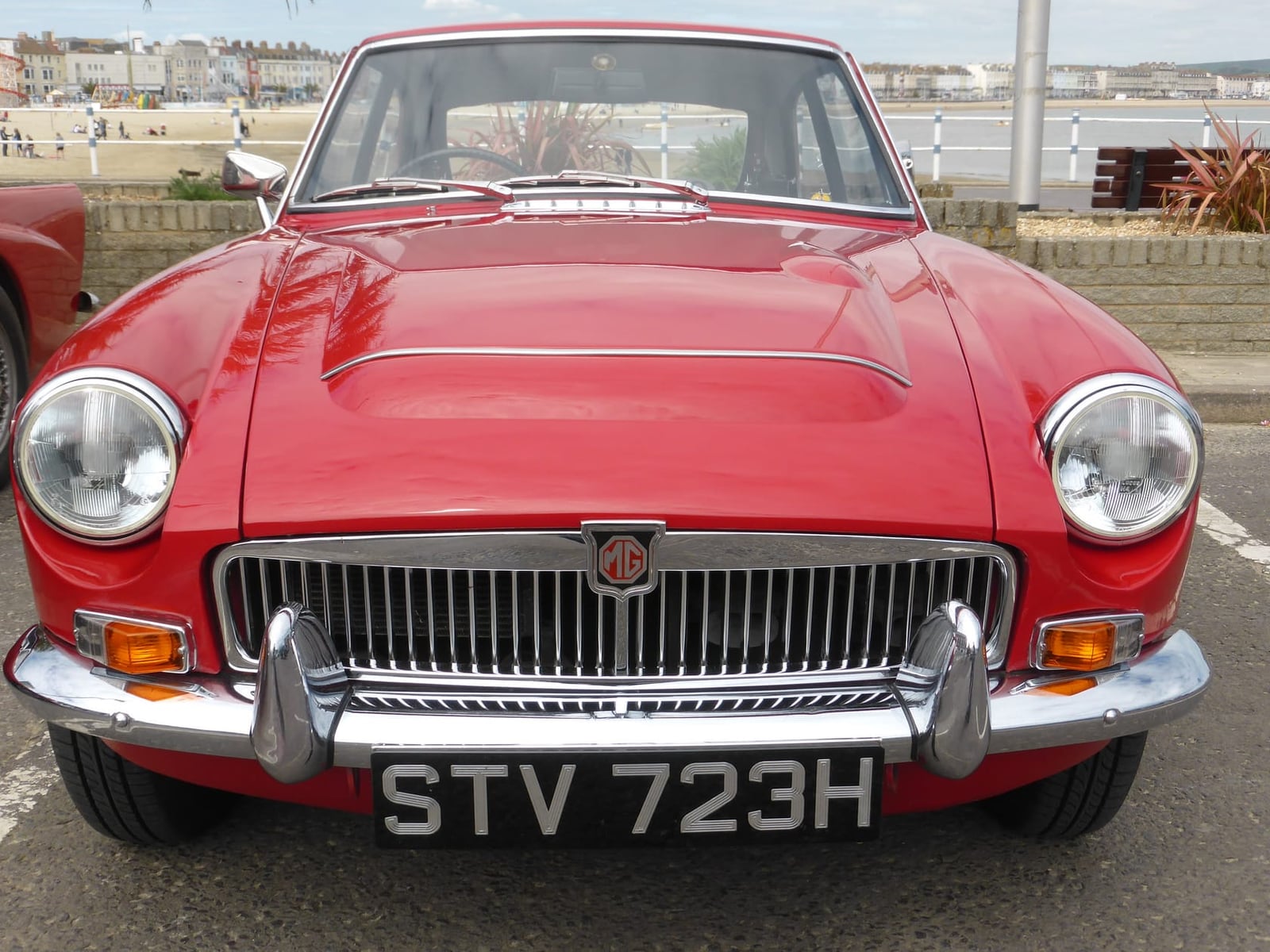 A red 1969 MGC GT with a UK registration, seen head-on beside the sea