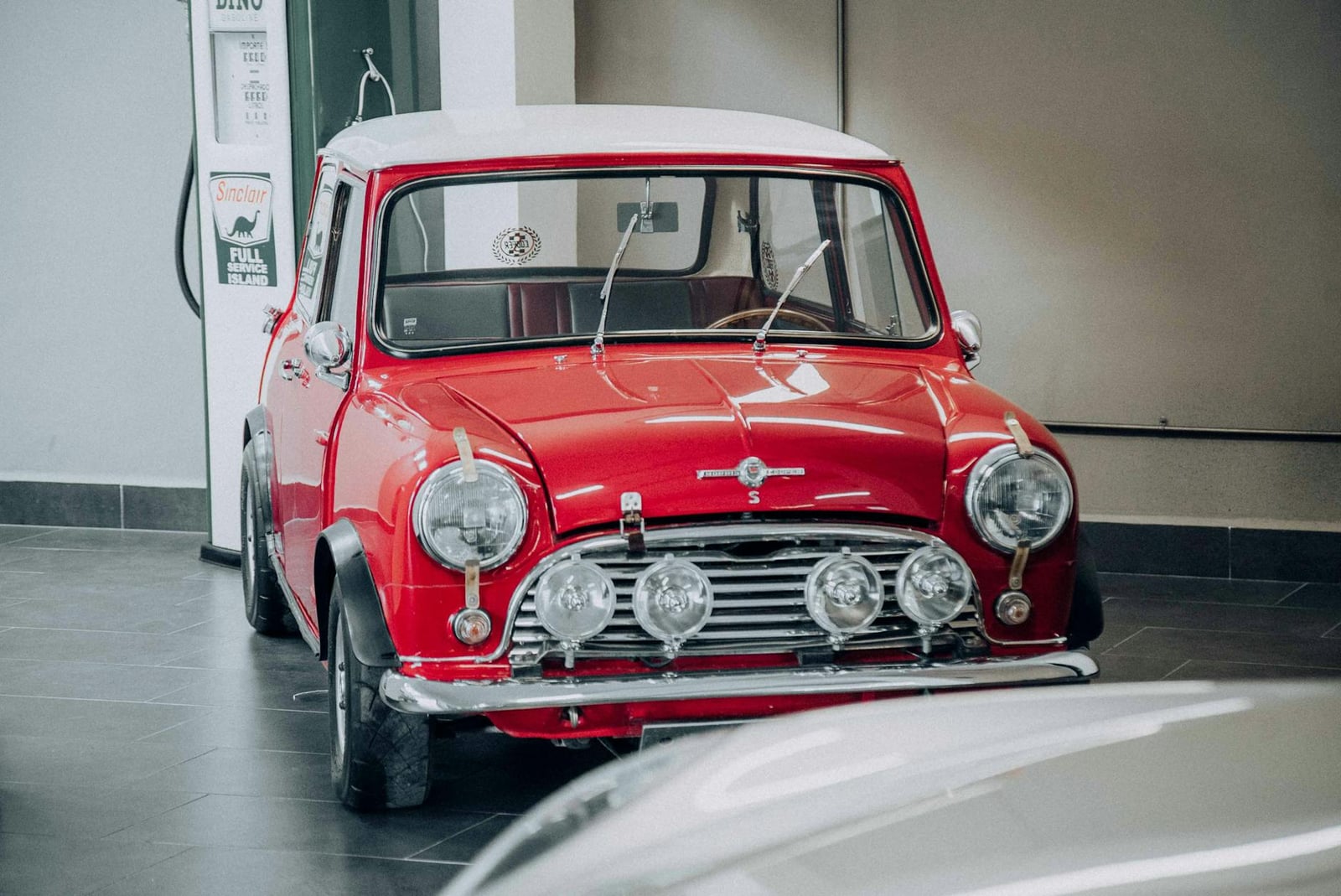 A red Mini Cooper S with a white roof and four auxiliary lamps, front three-quarter view indoors