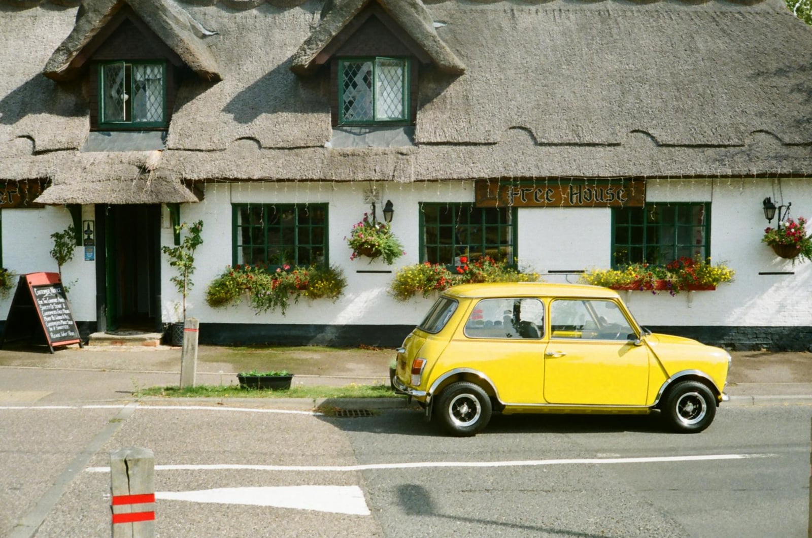 A yellow classic Mini with a white roof, parked in profile on a street outside a white thatched-roof pub hung with flower baskets