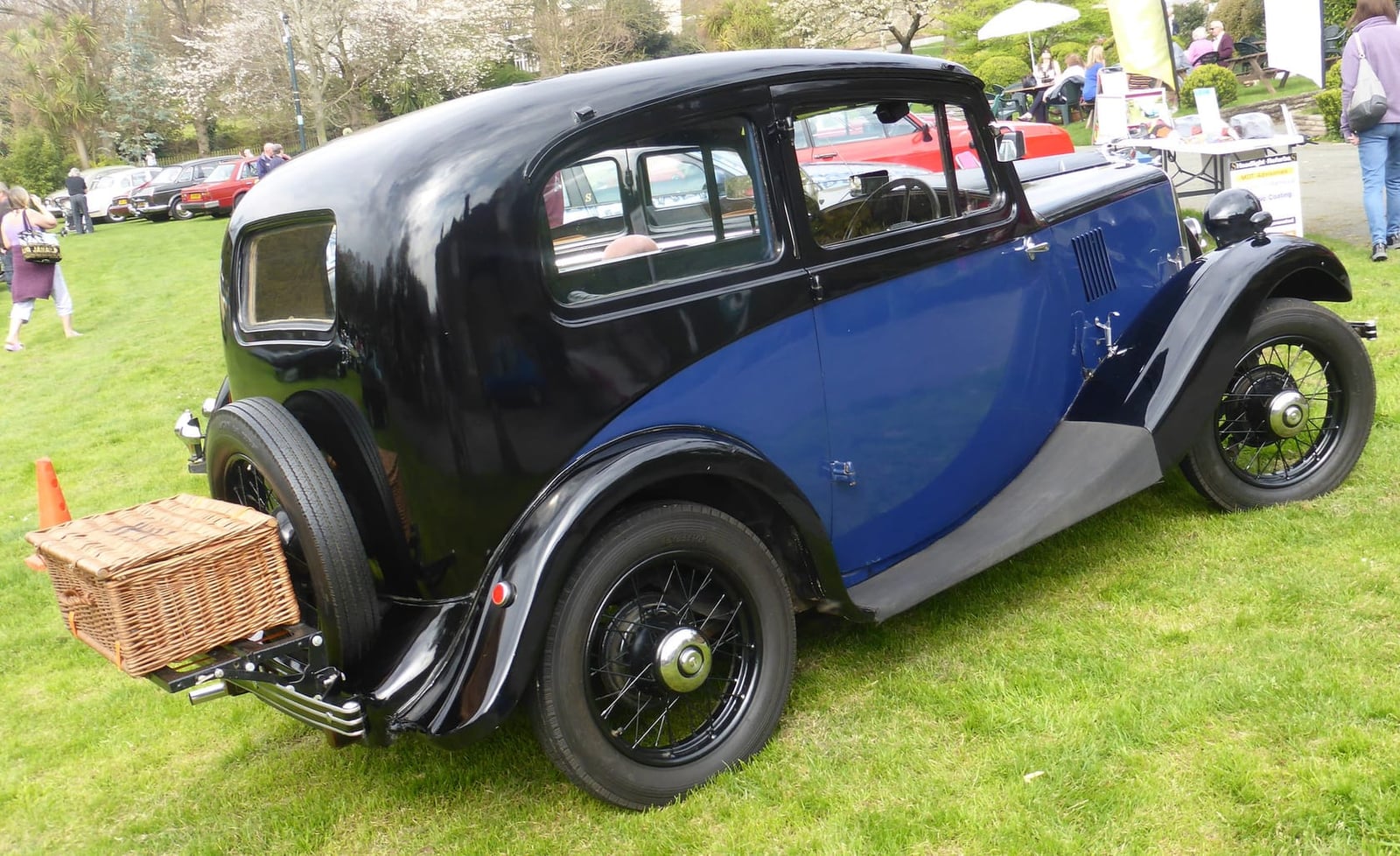 The rear three-quarter of a black and blue two-tone Morris Eight Series I on grass with a wicker basket on its luggage rack