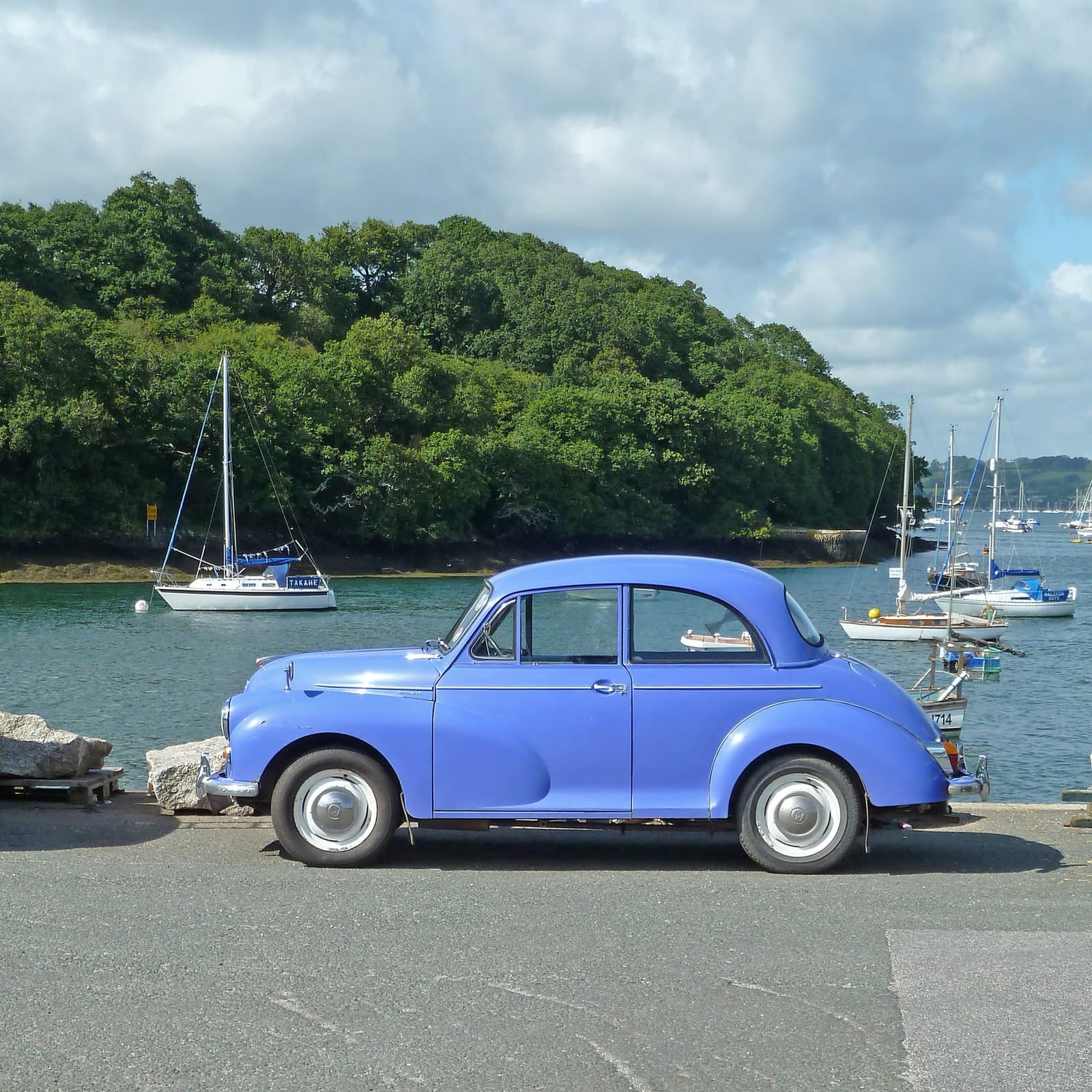 A blue Morris Minor 1000 parked on a Cornish quayside at Mylor with moored yachts behind