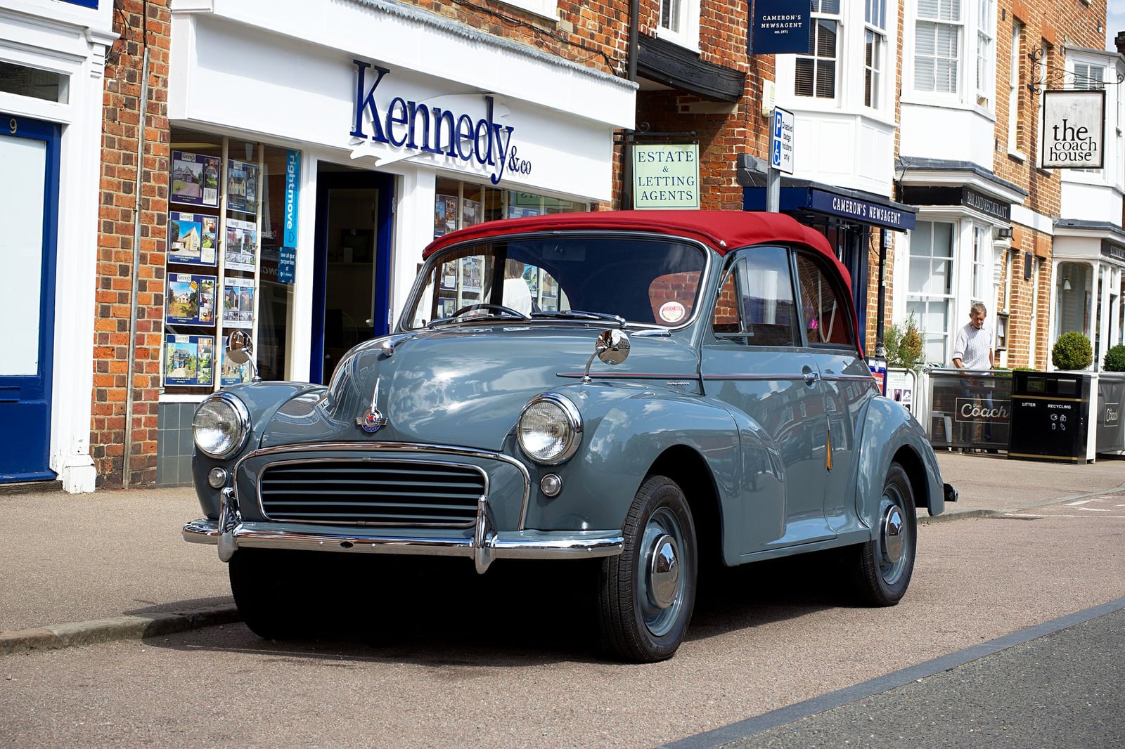 A grey Morris Minor convertible with a red soft top, front three-quarter view, parked on a British high street outside a row of shops
