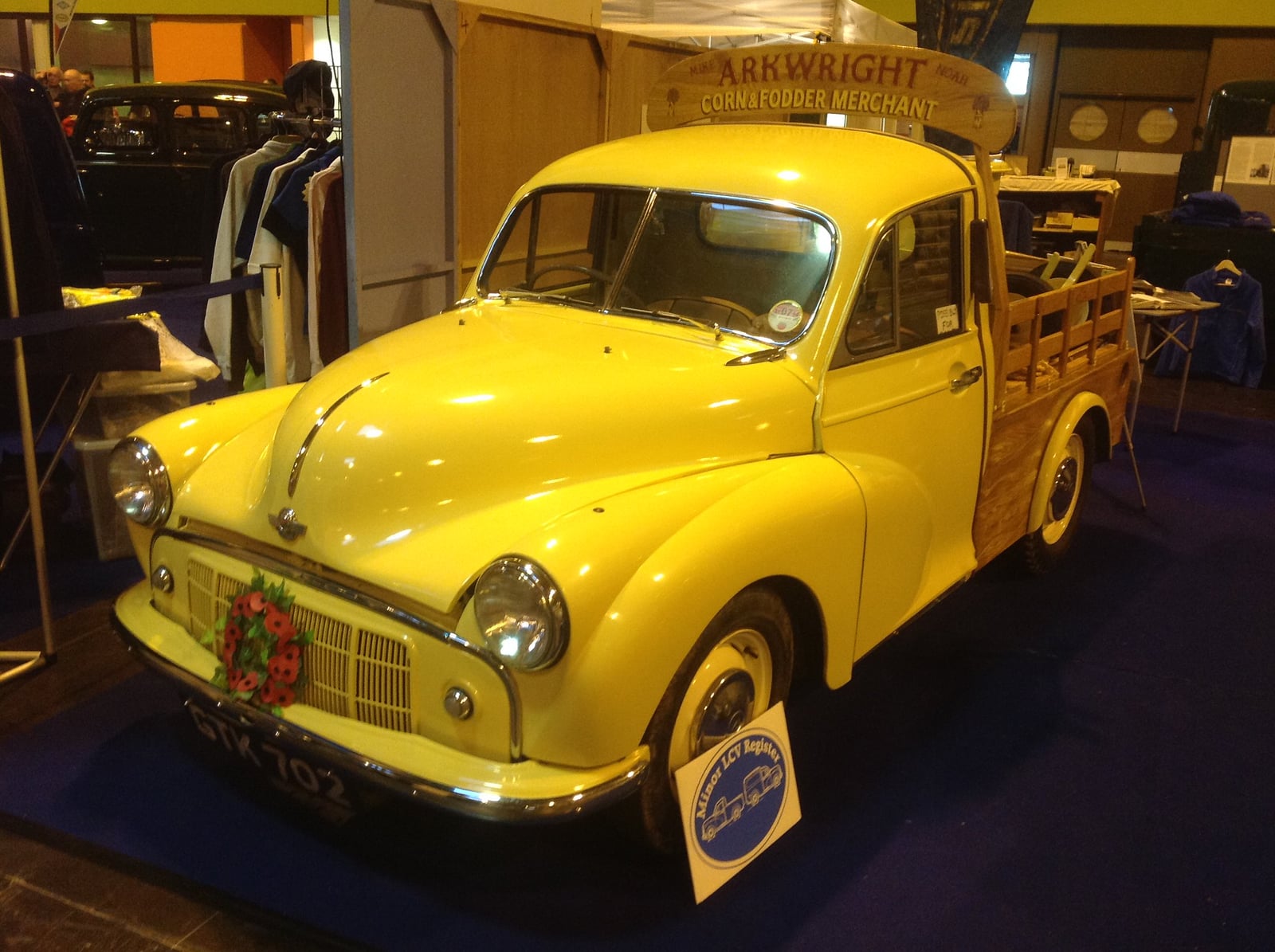 A yellow Morris Minor pick-up in period corn-merchant livery at an indoor show, front three-quarter view with a wood-railed load bed