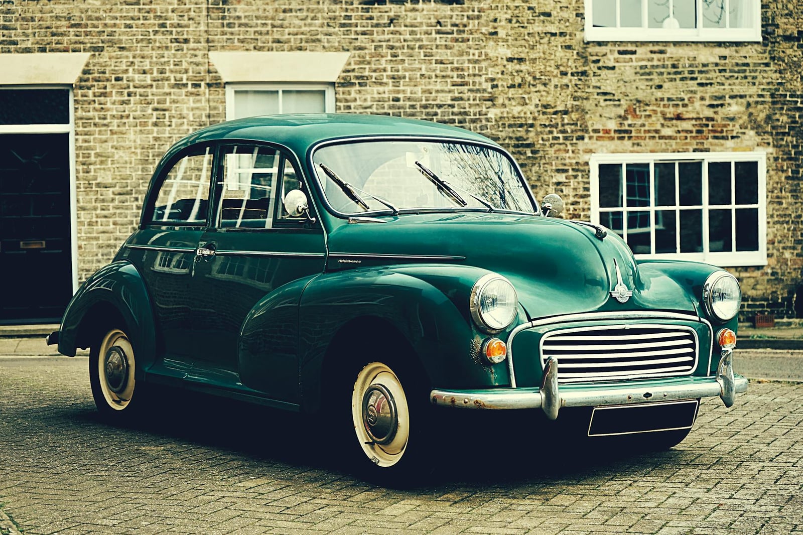 A dark green Morris Minor two-door saloon with cream wheels, three-quarter front view, parked on cobbles before a brick building