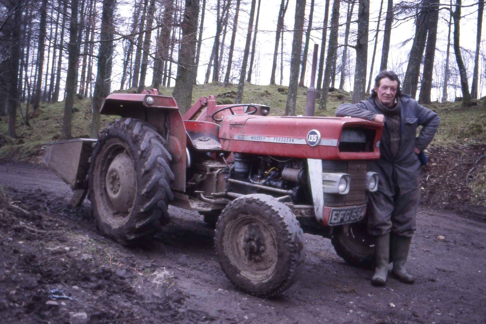 A muddy red Massey Ferguson 135 fitted with a front loader on a forestry track, its owner standing alongside in overalls