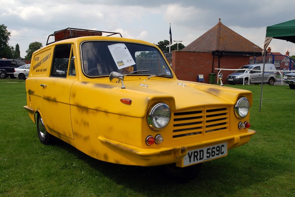 A yellow Reliant Regal Supervan III on grass at a classic vehicle show, front three-quarter view, UK registration YRD 569C