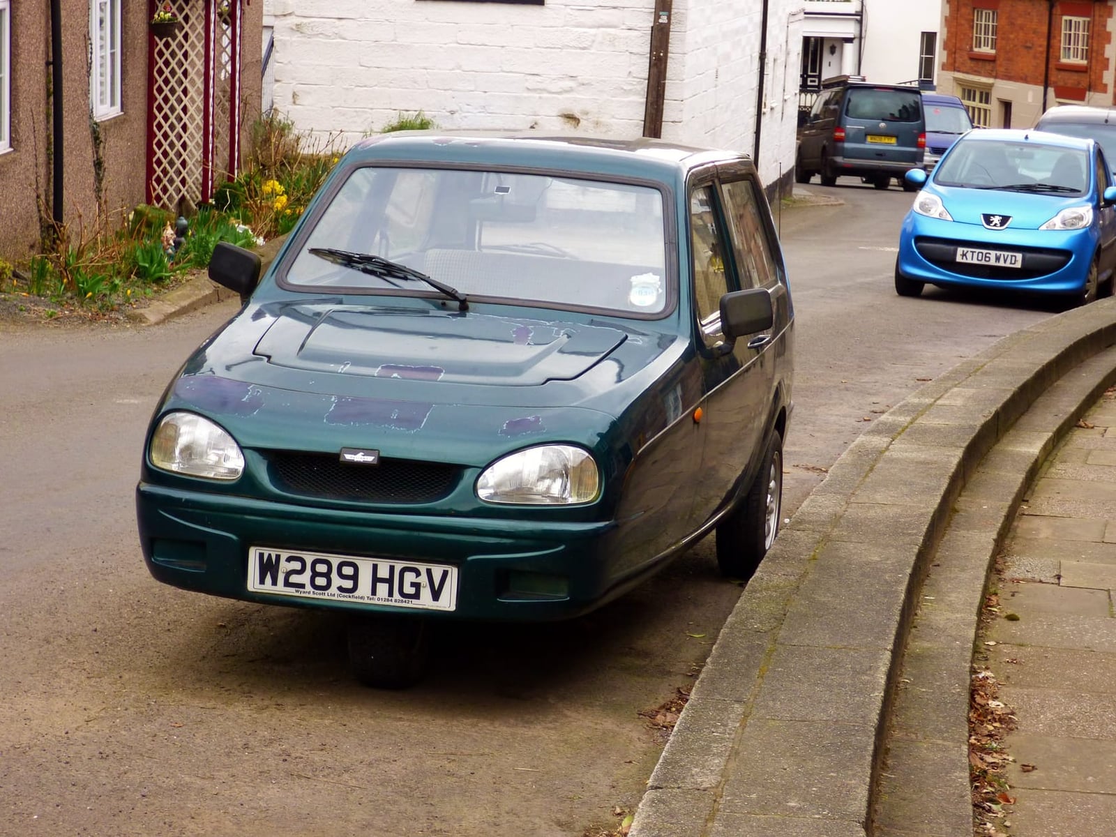A dark green late-model Reliant Robin parked on a village street, front three-quarter view, UK registration W289 HGV