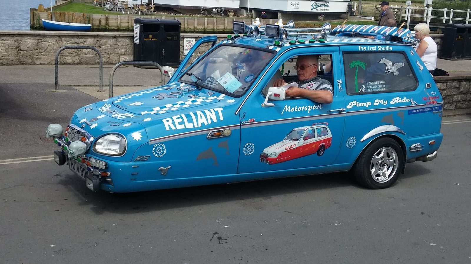 A blue Reliant Robin covered in rally decals and fitted with extra lamps, being driven along a quayside