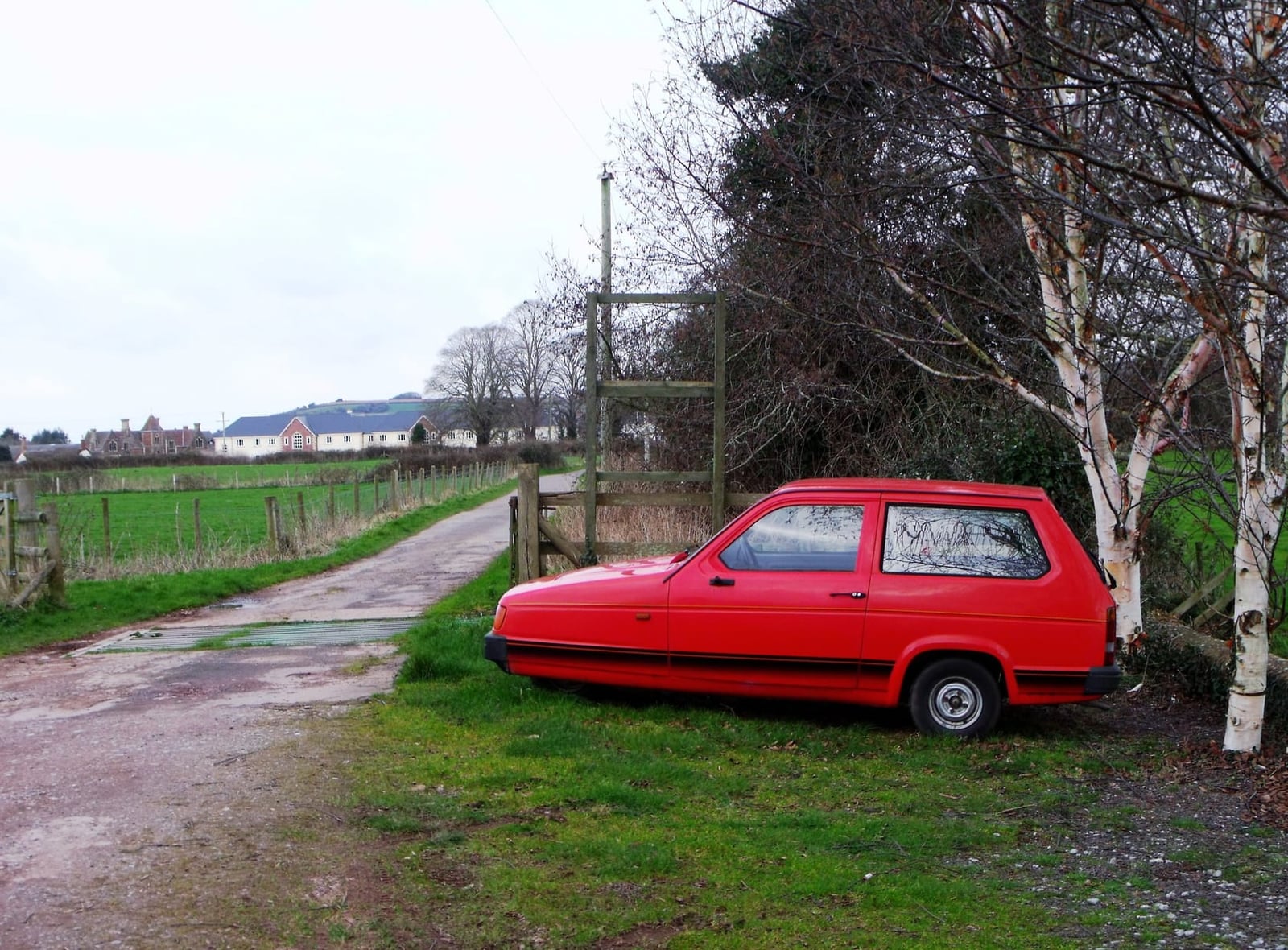 A red Reliant Robin parked on a rural lane beside bare trees, side view