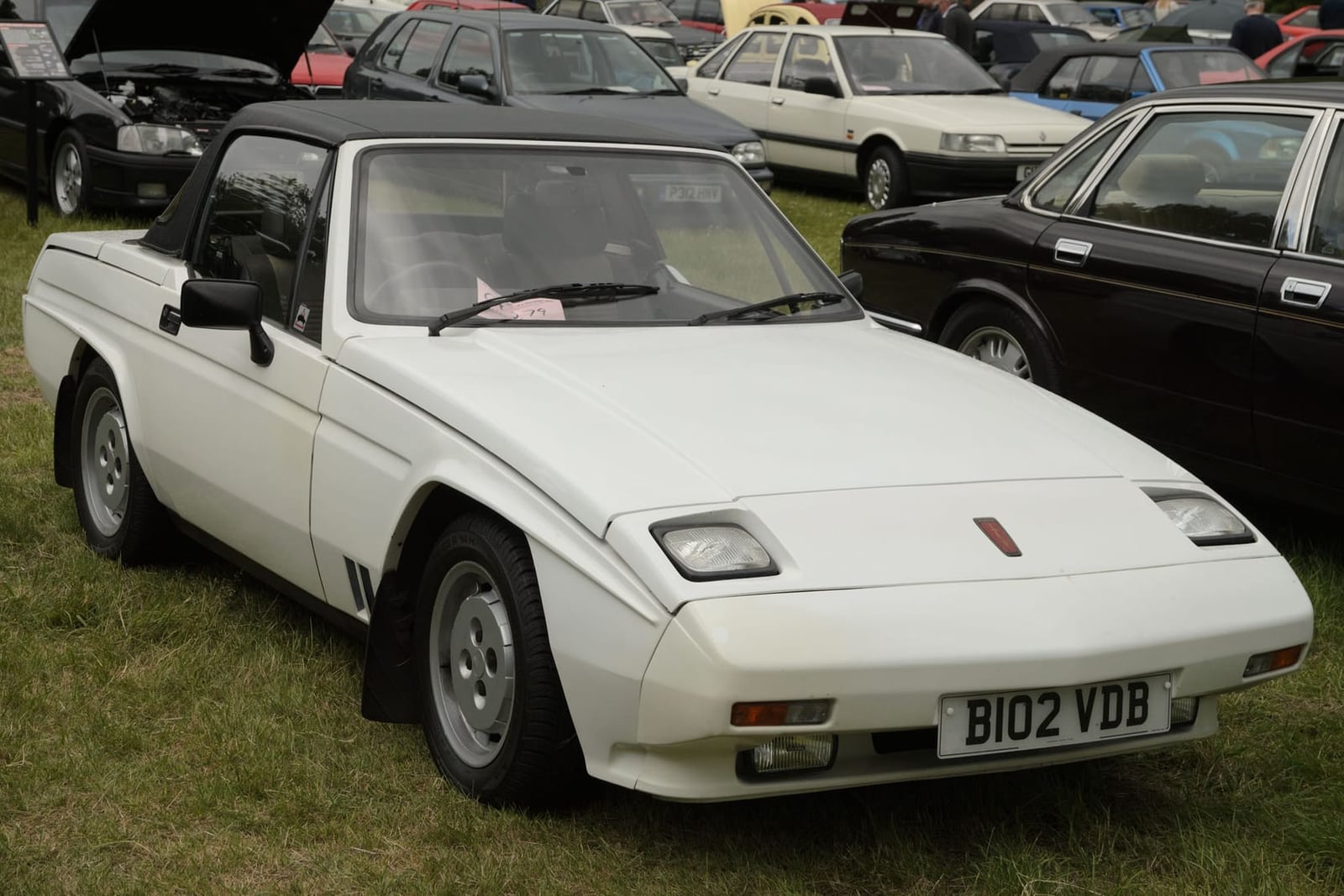 A white Reliant Scimitar SS1 1300 on grass at a classic car show, front three-quarter view, UK registration B102 VDB