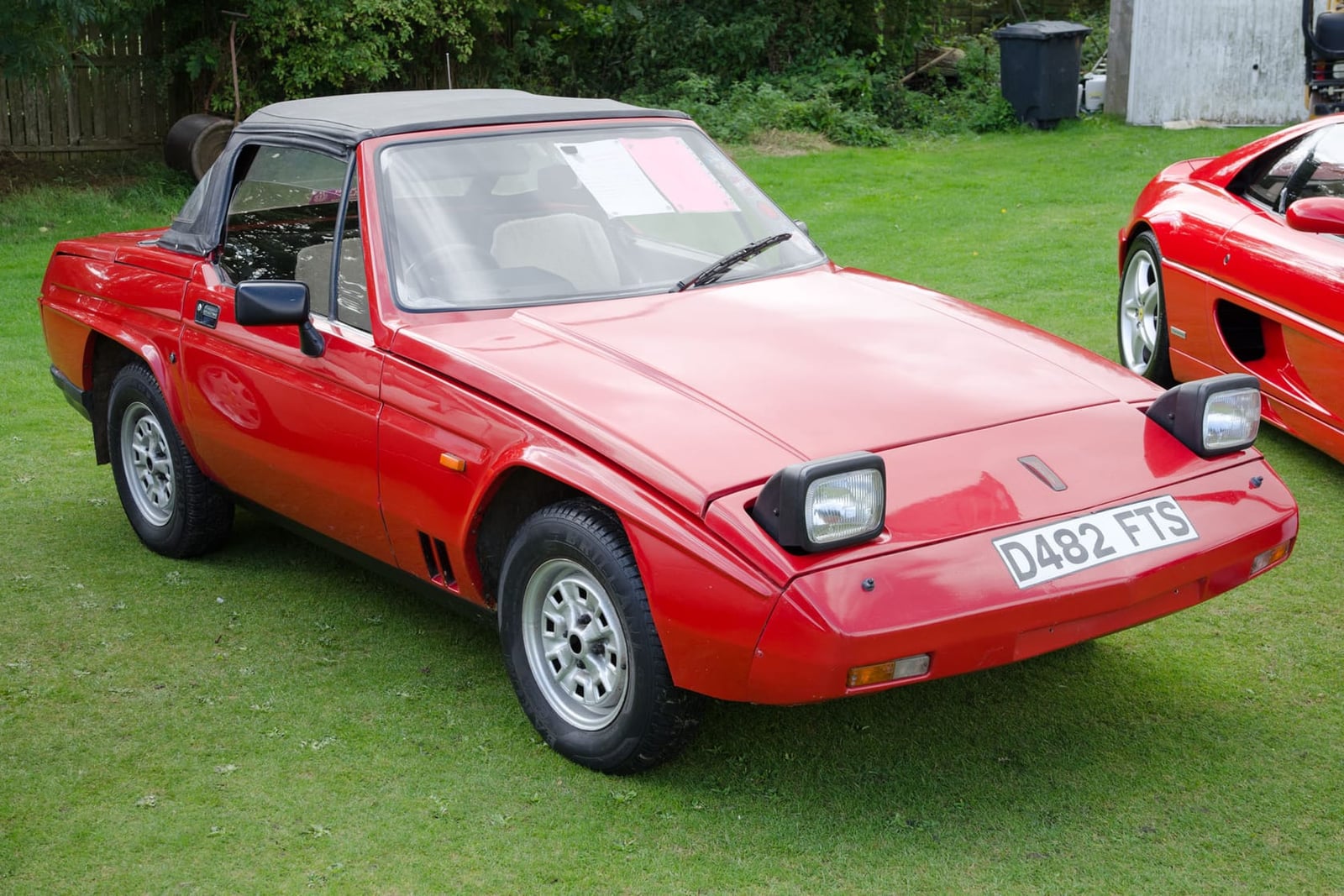 A red 1987 Reliant Scimitar SS1 on grass at a classic car show, front three-quarter view, UK registration D482 FTS