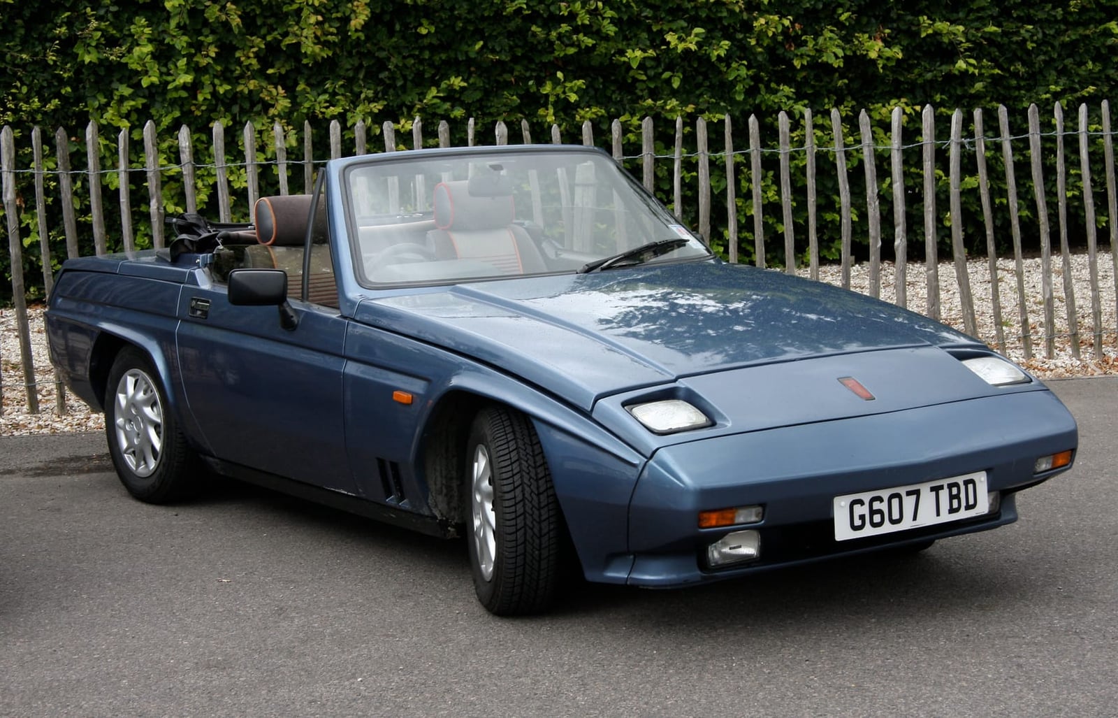 A dark blue Reliant Scimitar SS1 with the hood down parked beside a fence, side view, UK registration G607 TBD