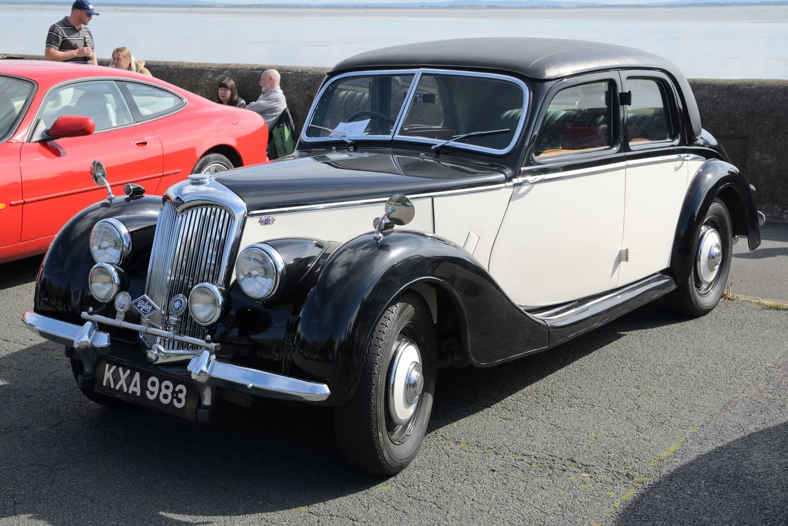A black and cream two-tone 1949 Riley RMA saloon parked by a harbour wall, front three-quarter view, UK registration KXA 983