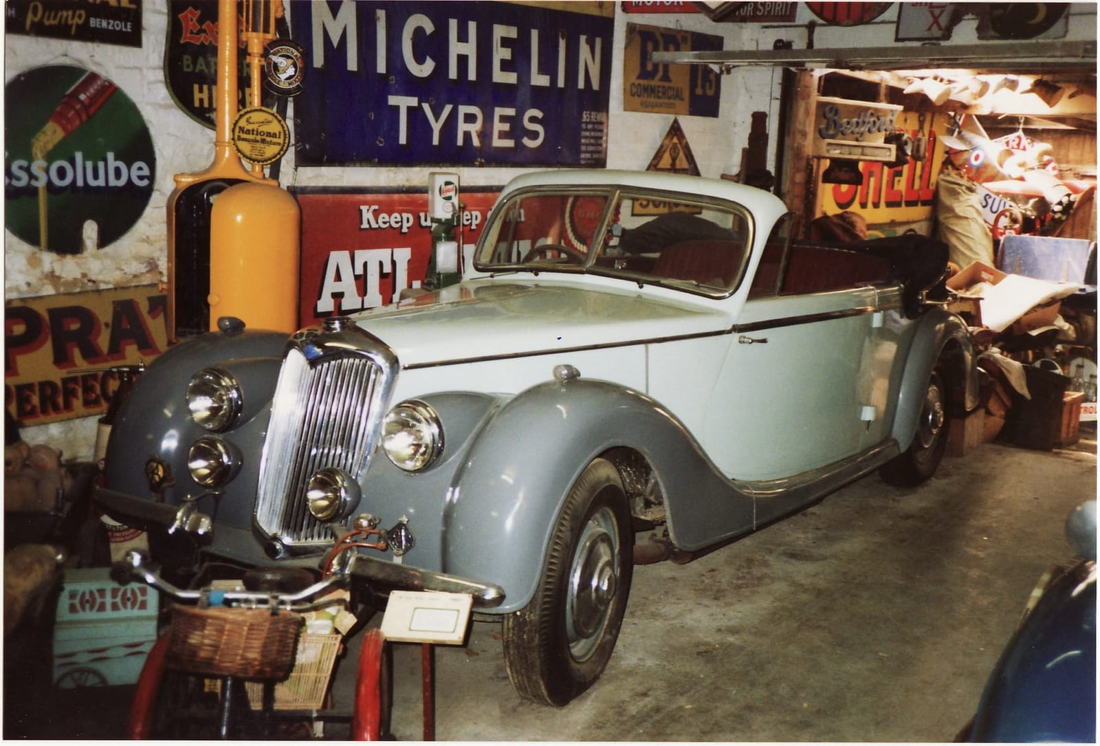A grey and blue Riley RMD drophead in a cluttered workshop surrounded by vintage garage signs