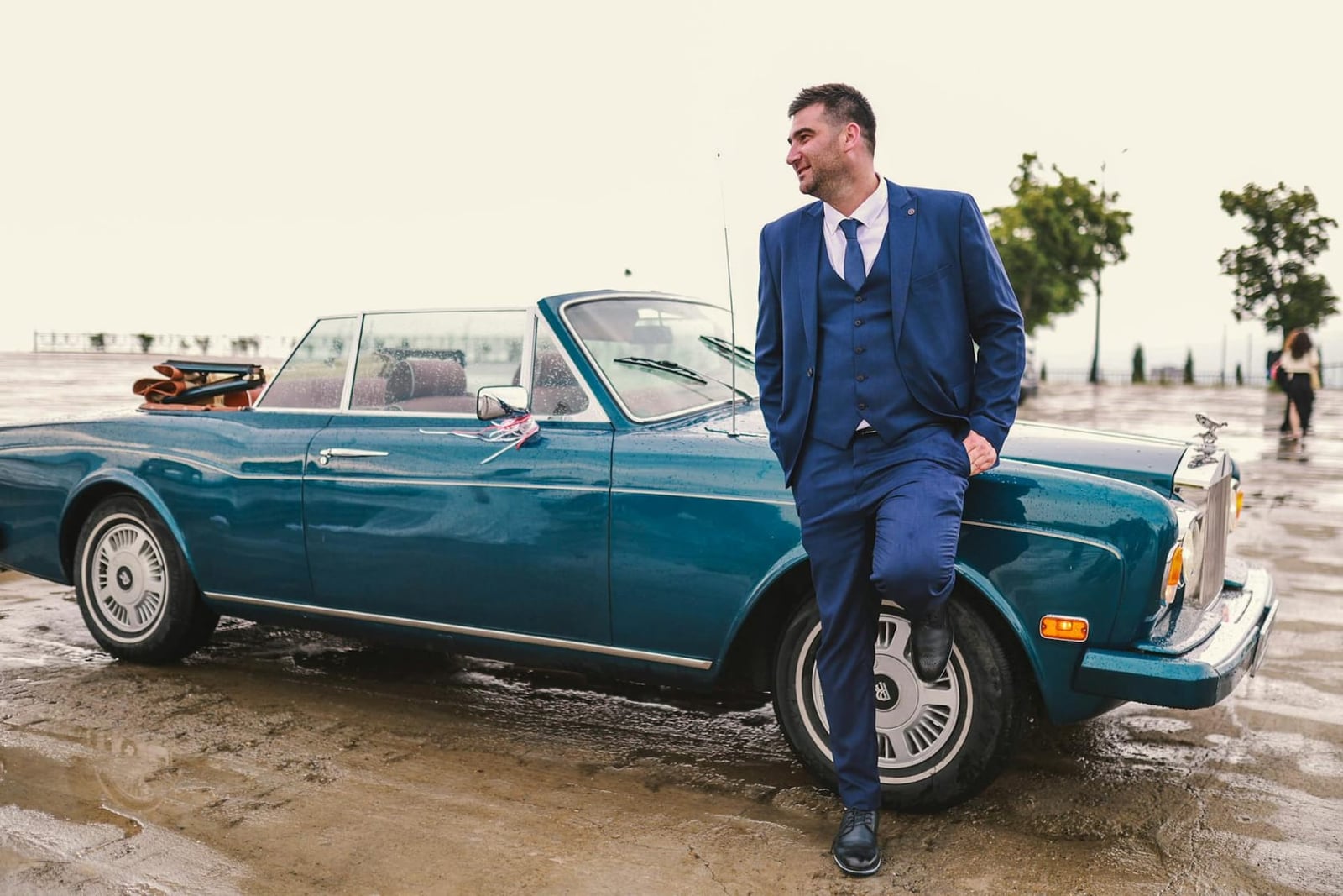 A teal Rolls-Royce Corniche convertible with a groom leaning against it by the seafront