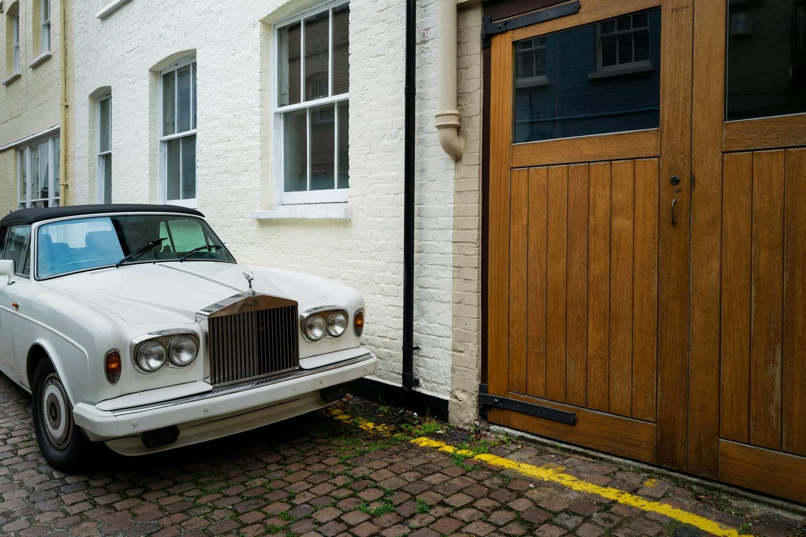 A white Rolls-Royce Corniche convertible parked beside a wooden garage door in a mews