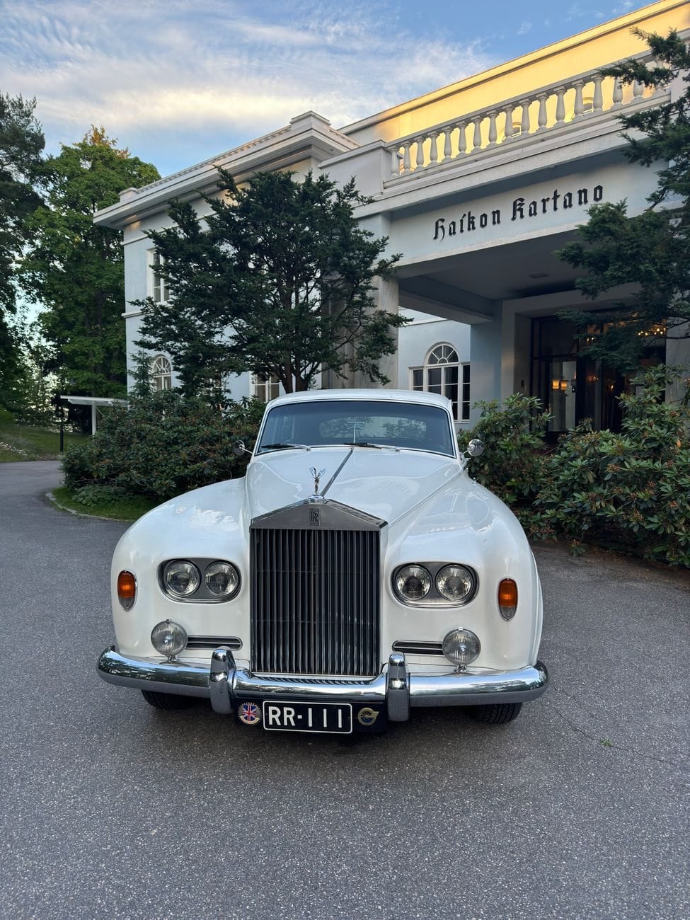 A white Rolls-Royce Silver Cloud III parked outside a manor house