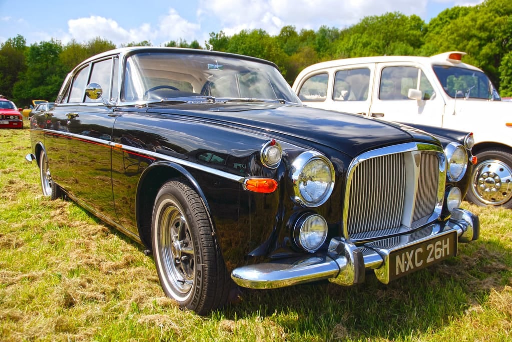 A dark blue Rover P5B coupe on grass at a classic vehicle show, front three-quarter view, UK registration NXC 26H