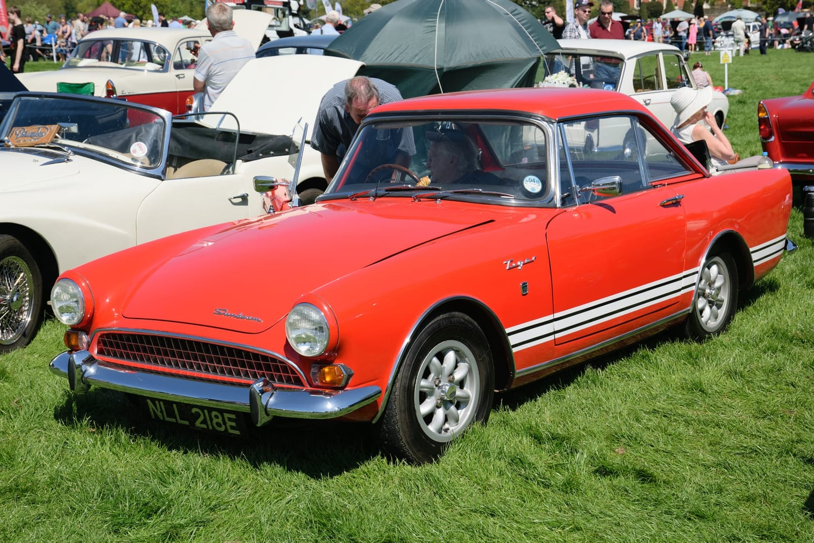 A red Sunbeam Tiger with white side stripes at a busy showfield, front three-quarter view