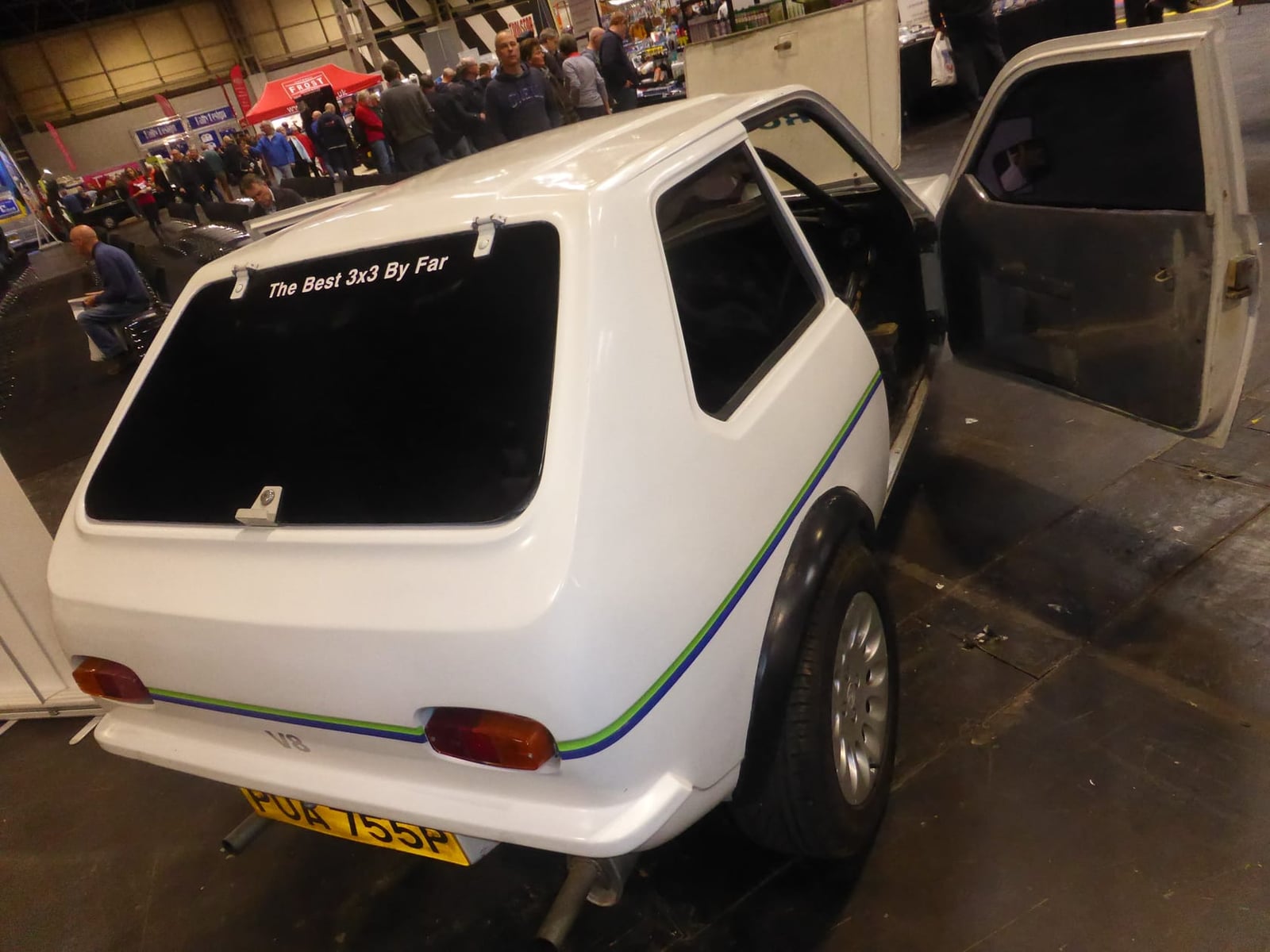 A white Reliant Robin with its rear hatch raised, showing the load space, at an indoor show