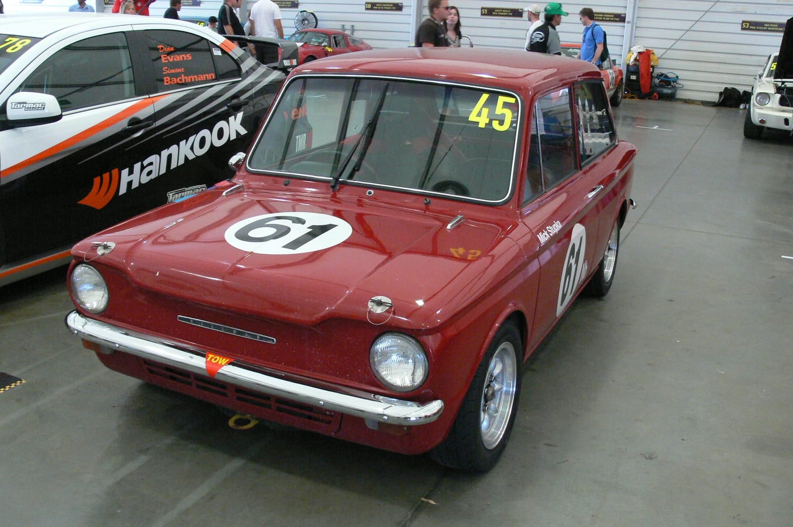 A red Hillman Imp saloon prepared for racing, carrying number 61 and sponsor decals, in a paddock
