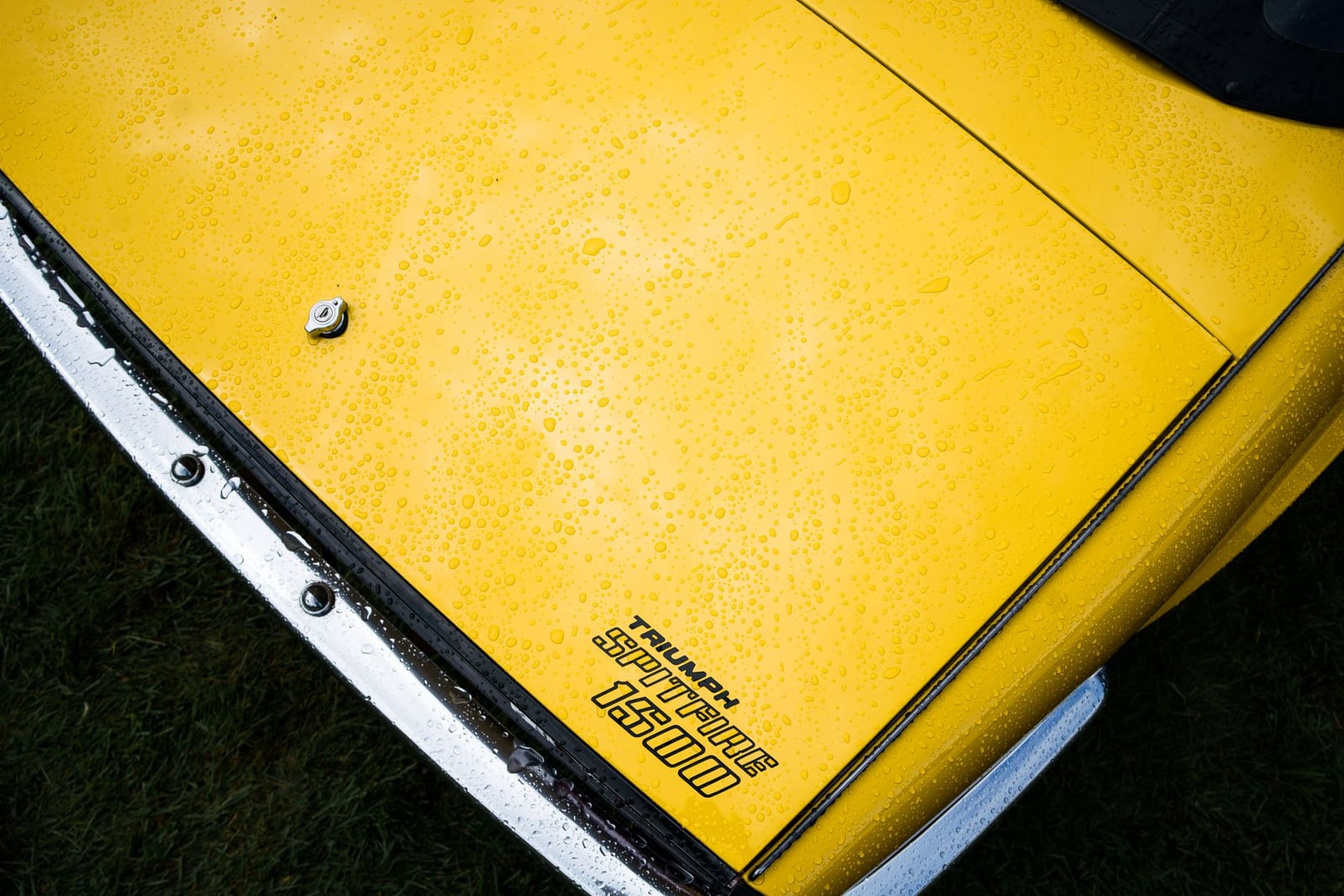 Close-up of a yellow Triumph Spitfire 1500 bonnet beaded with rain, showing the chrome script badge