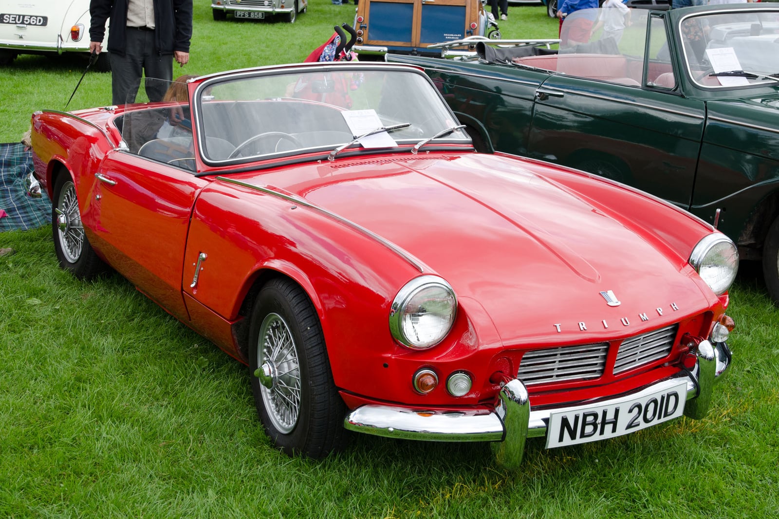 A red Triumph Spitfire Mk2 with wire wheels, top down, front three-quarter view on grass at a show