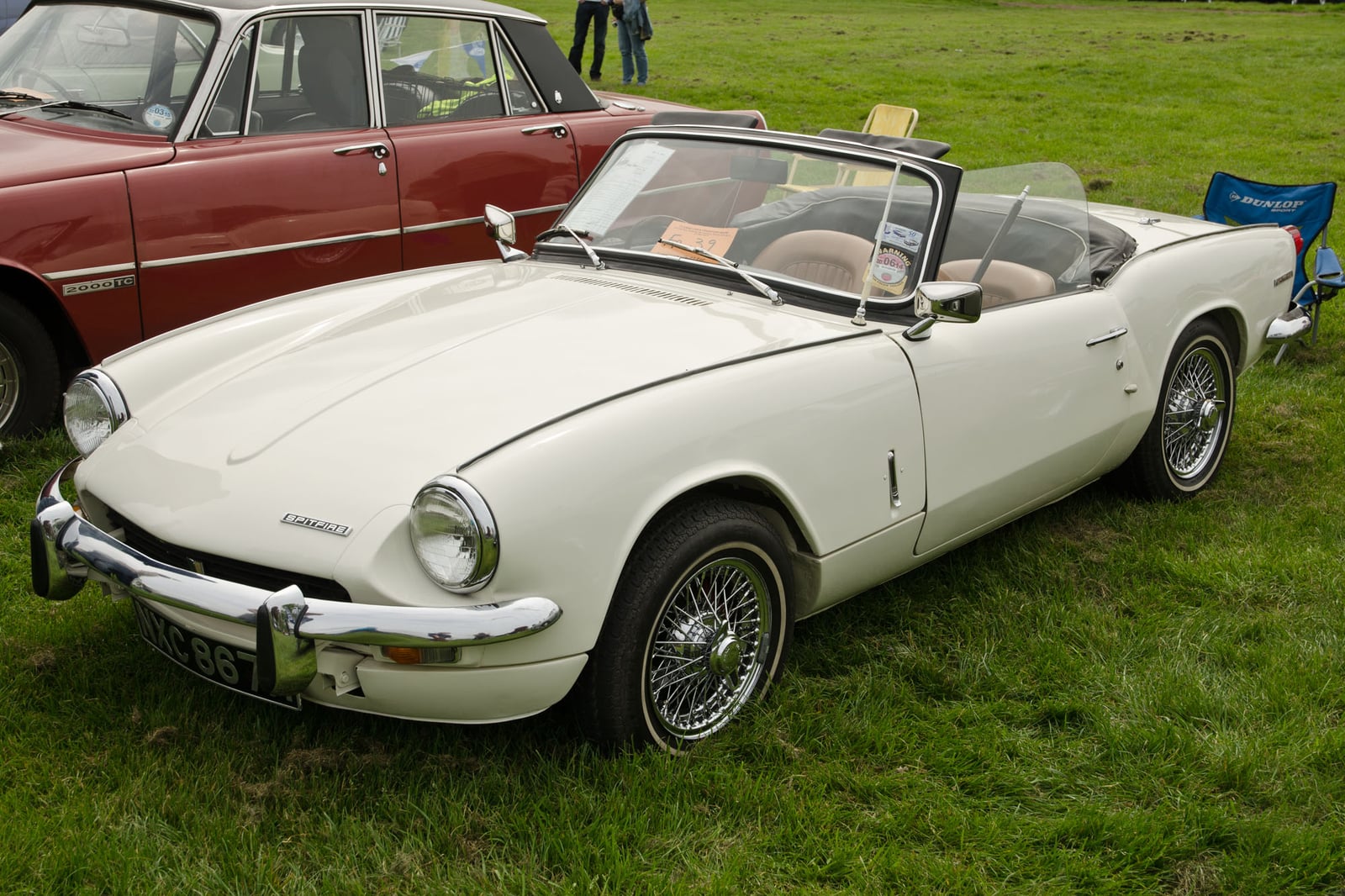A cream Triumph Spitfire Mk3 with wire wheels, top down, front three-quarter view on grass