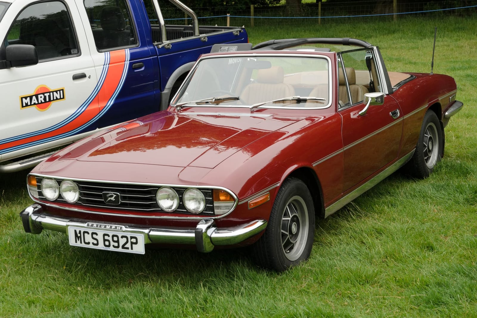 A maroon 1976 Triumph Stag Mk2 with its hood down and a UK registration on a show field
