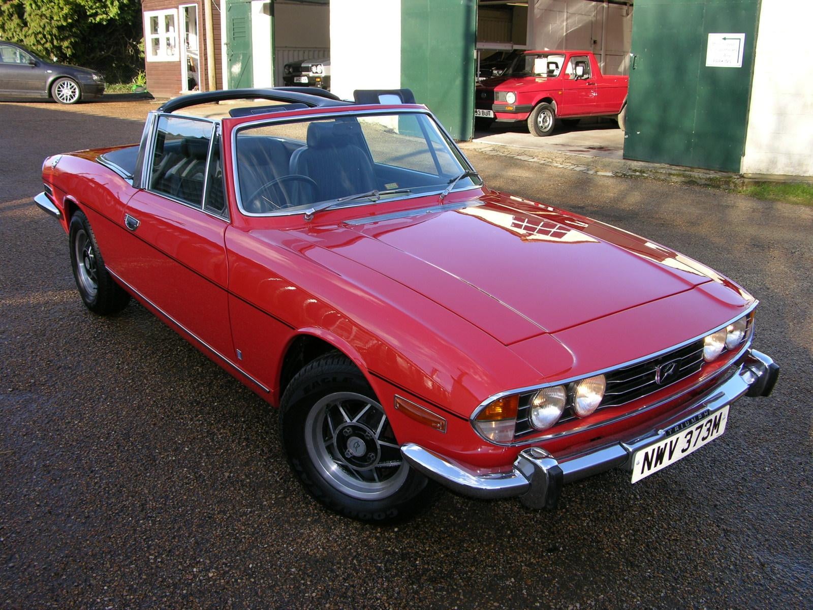 A red Triumph Stag convertible with the top down, showing the T-bar roll hoop
