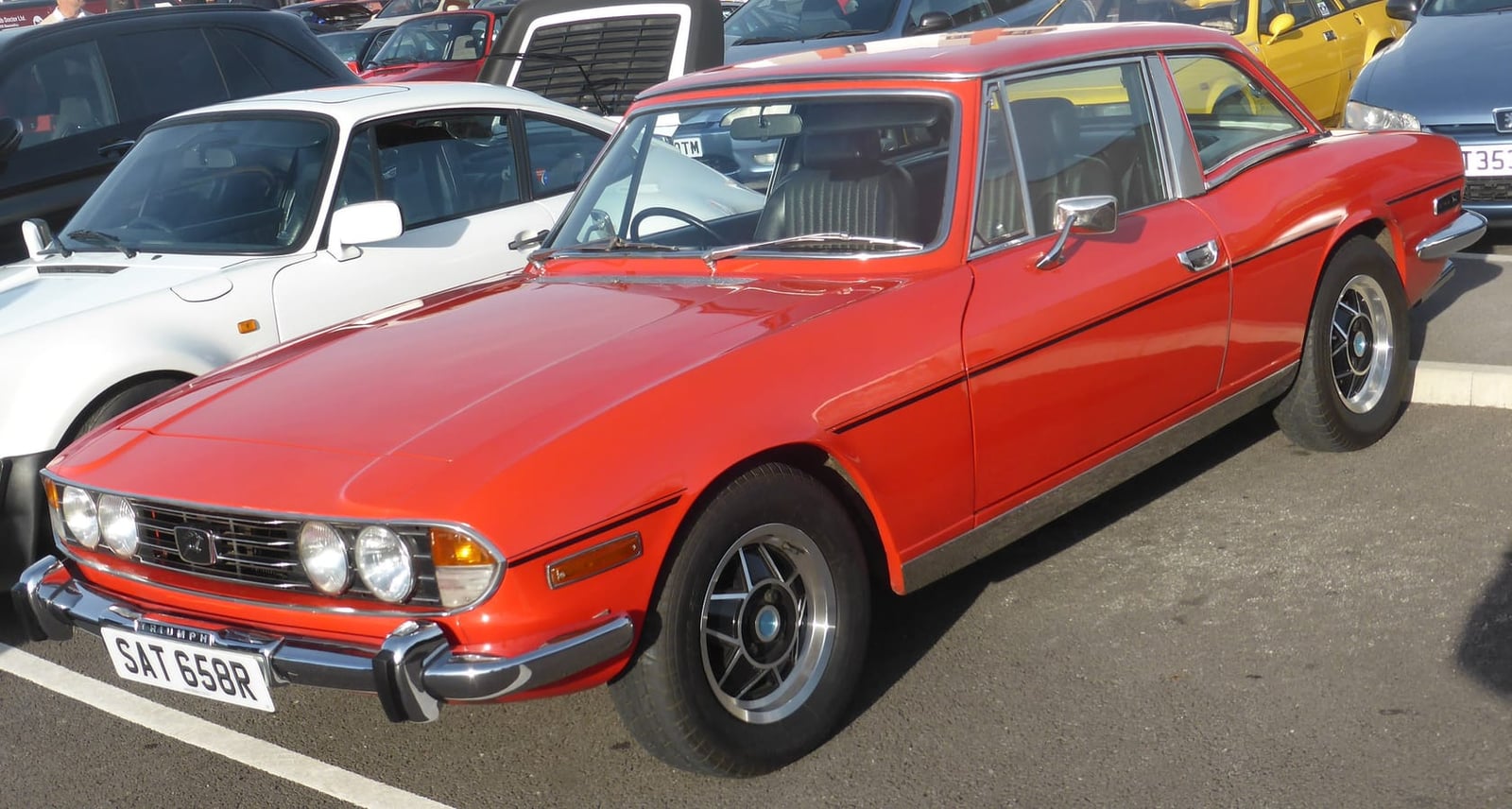 A red 1977 Triumph Stag with its factory hardtop fitted and a UK registration in a car park at a show