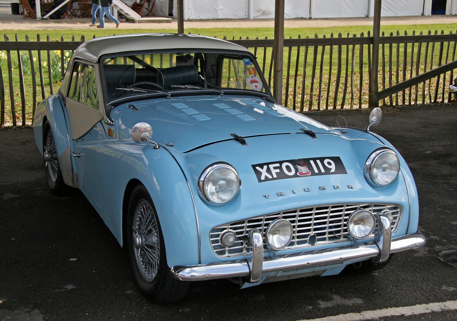 A pale blue Triumph TR3 with a hardtop, front three-quarter view, by a fence at a show