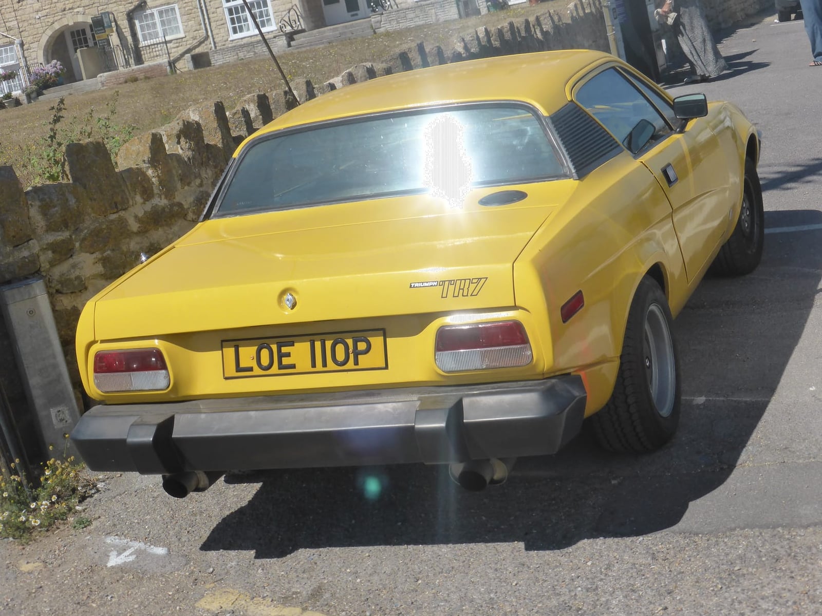 A yellow 1976 Triumph TR7 coupe parked beside a stone wall, rear three-quarter view, UK registration LOE 110P