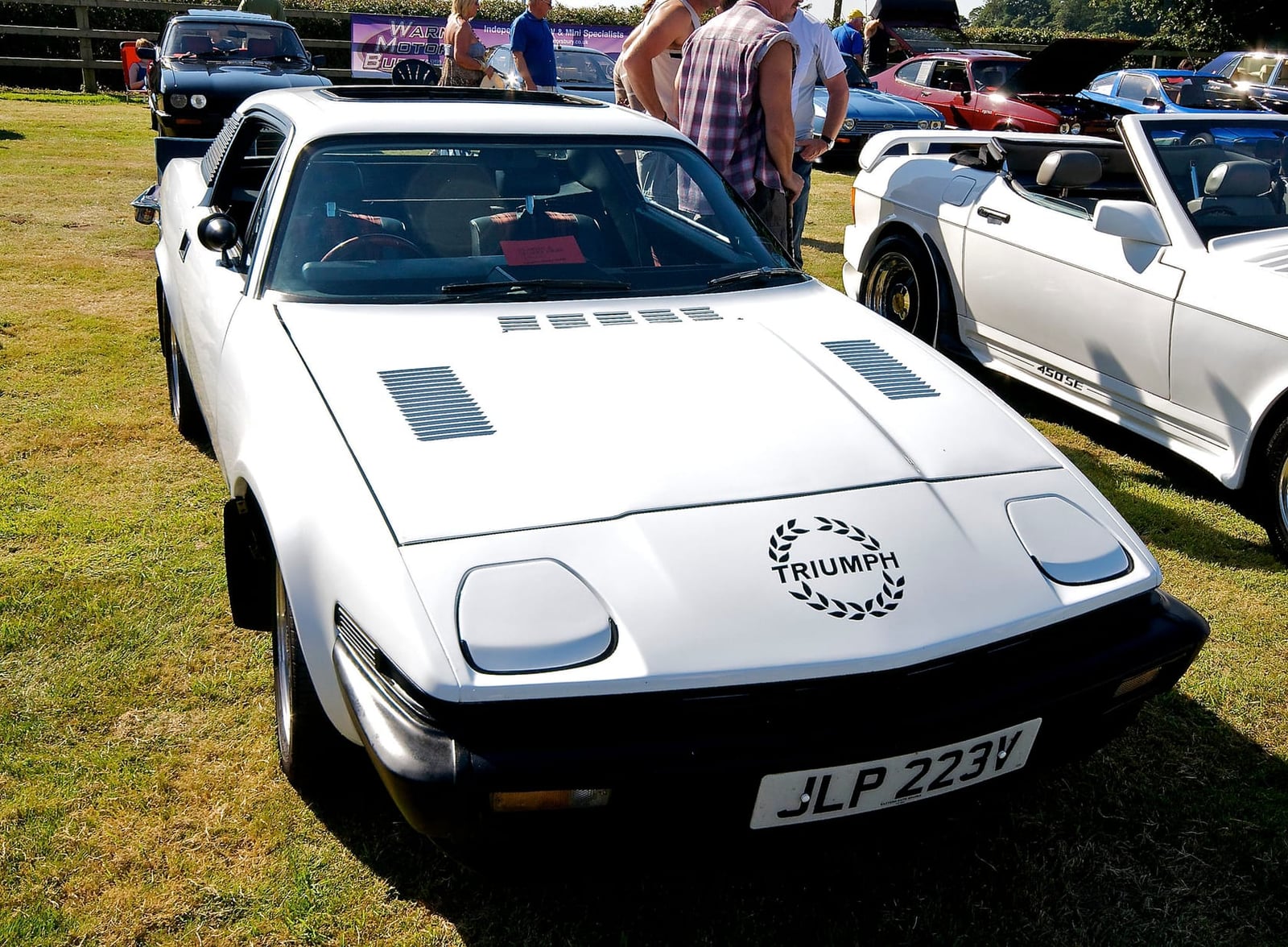 A white Triumph TR7 with Triumph laurel decals at a show, other TR7s behind