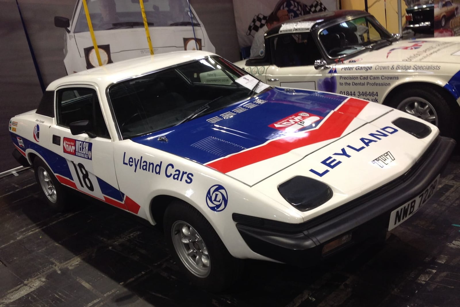 A white 1976 works Triumph TR7 rally car in red and blue Leyland Cars livery, race number 18, on an indoor display stand, UK registration NWB 722P