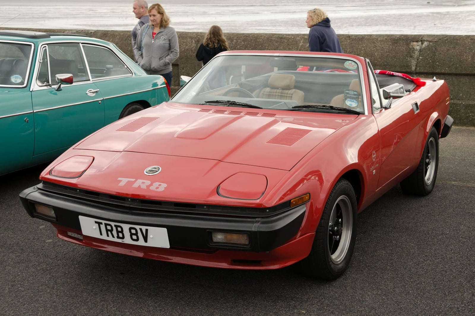 A red 1981 Triumph TR8 convertible parked at a seafront, front three-quarter view, registration TRB 8V